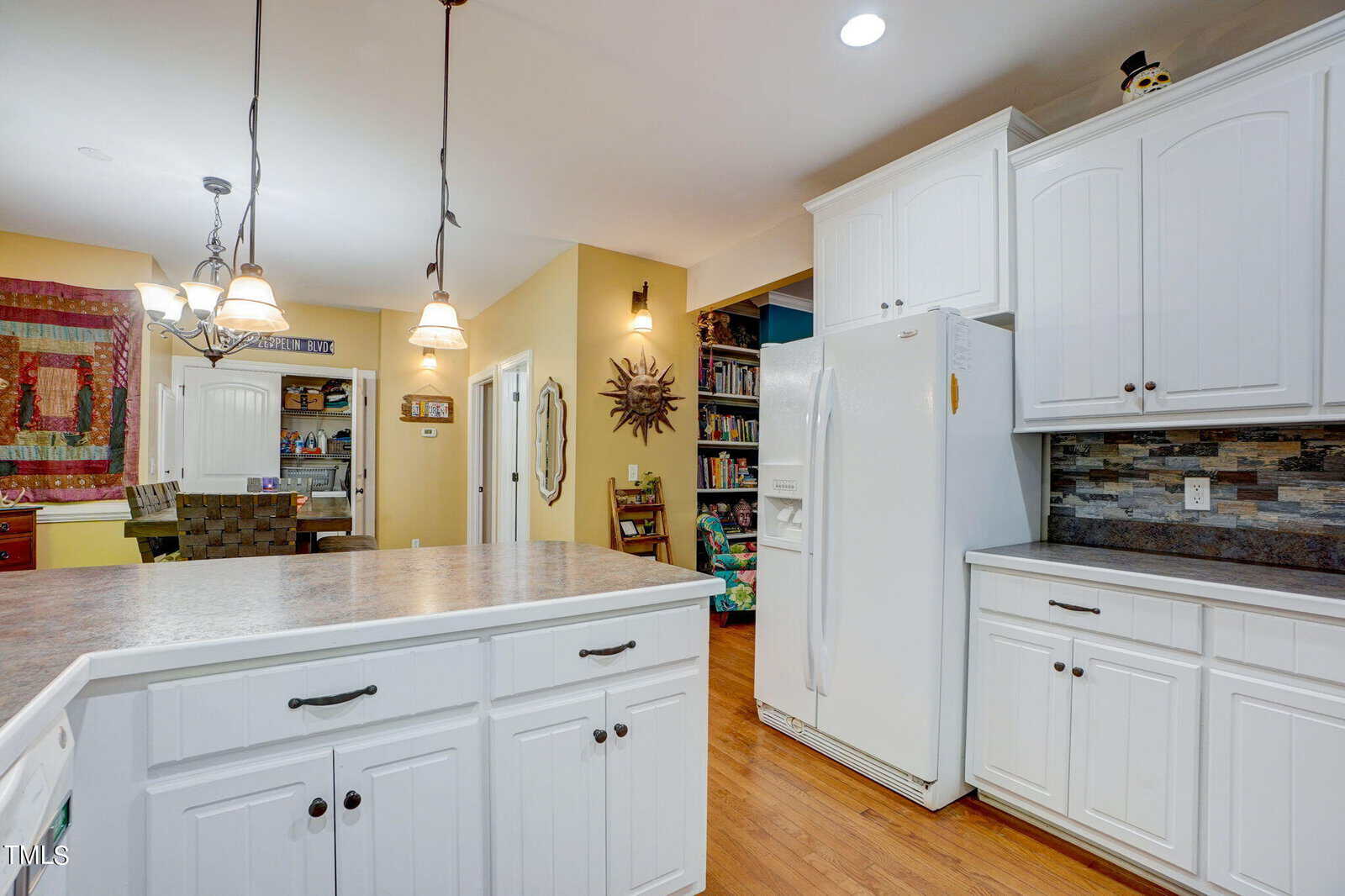 A kitchen with white cabinets, a white refrigerator, and a large island with a stone countertop. Pendant lights hang above the island. A dining area and bookshelf are visible in the background.