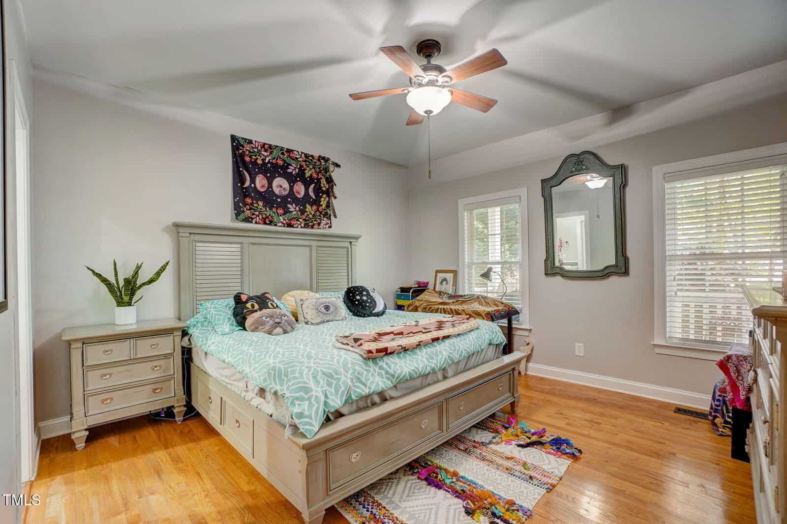 A cozy bedroom with a light wood bed, colorful pillows, a tapestry above the headboard, a ceiling fan, a large mirror, and hardwood floors with vibrant rugs. Natural light enters through two windows with blinds.