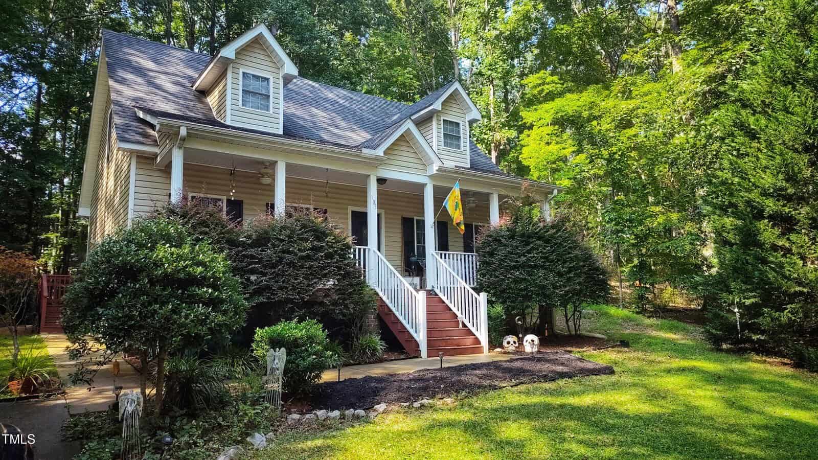 A beige two-story house with a front porch, white railings, two dormer windows, and a yellow flag. The house is surrounded by bushes, trees, and a green lawn under bright sunlight.
