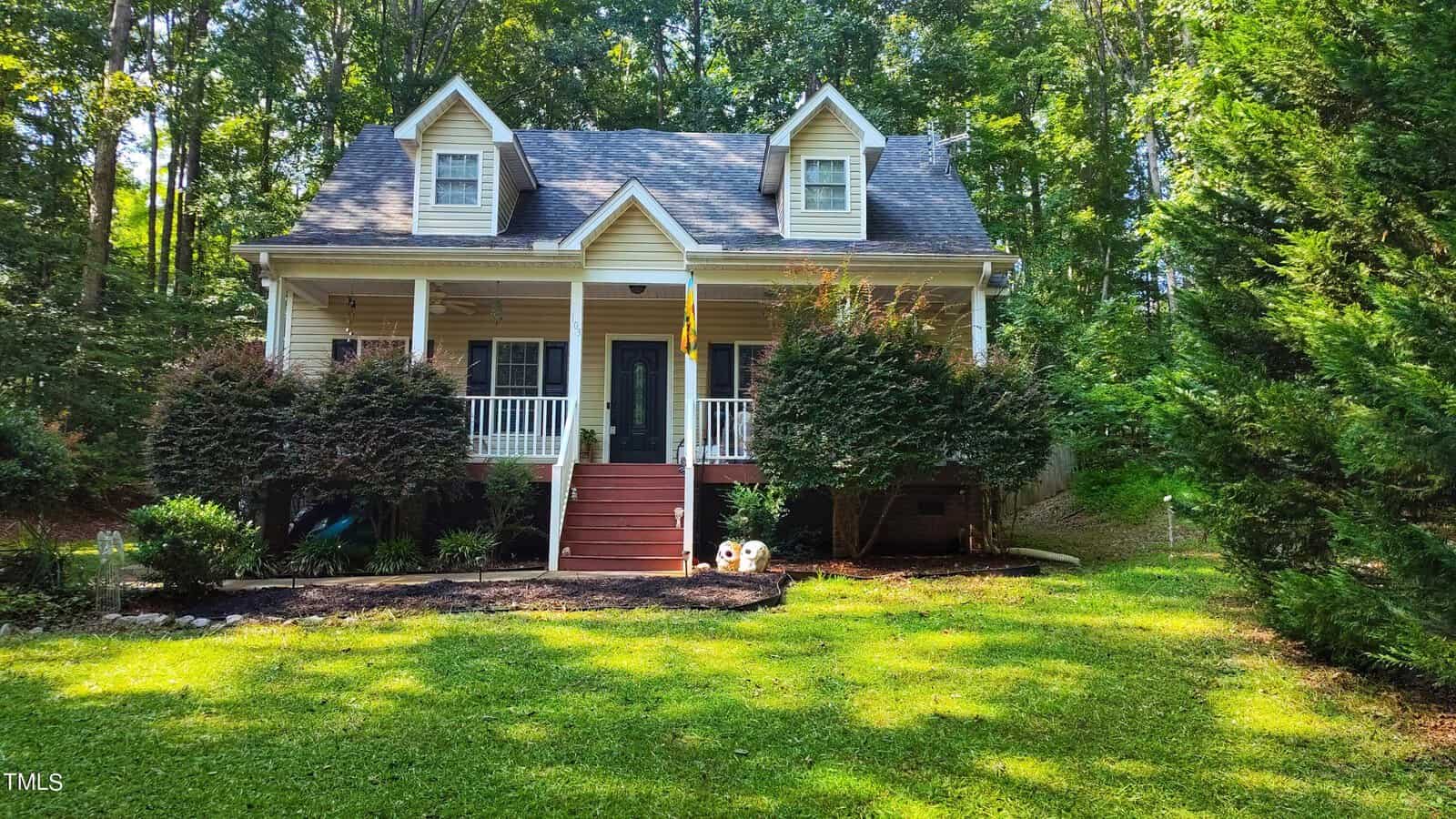 A two-story house with a covered front porch, two dormer windows, and red stairs leading to the entrance. The yard is grassy with shrubs and trees, and a wooded area is visible in the background.