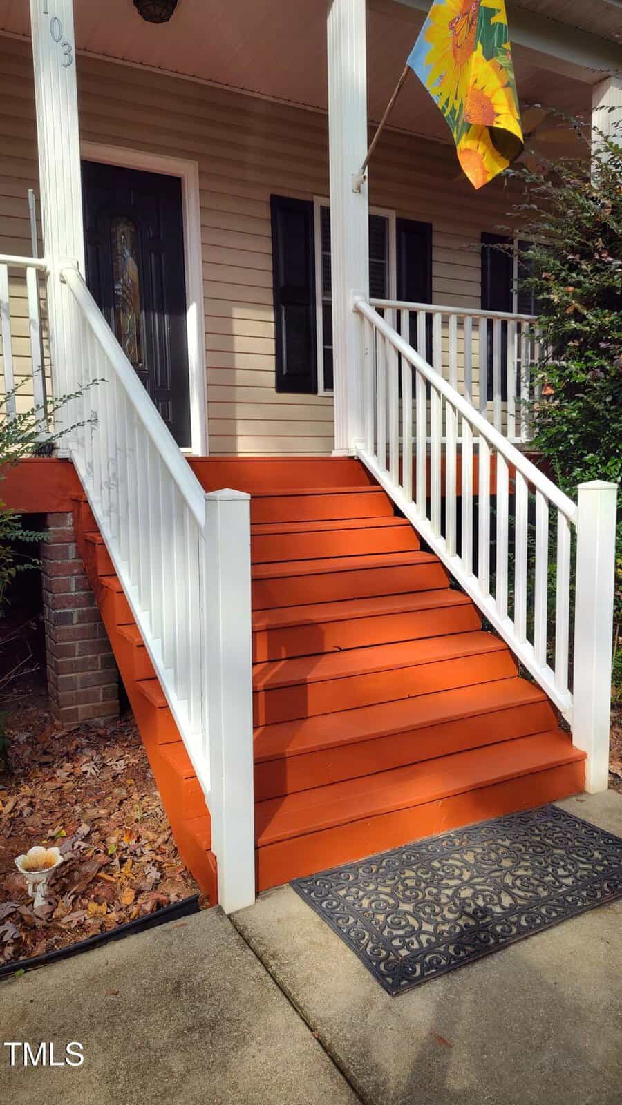 Front porch steps painted orange with white railings lead up to a door with glass panels. A doormat lies at the base of the steps, and a colorful flag hangs near the porch. Shrubs and fallen leaves are visible beside the steps.