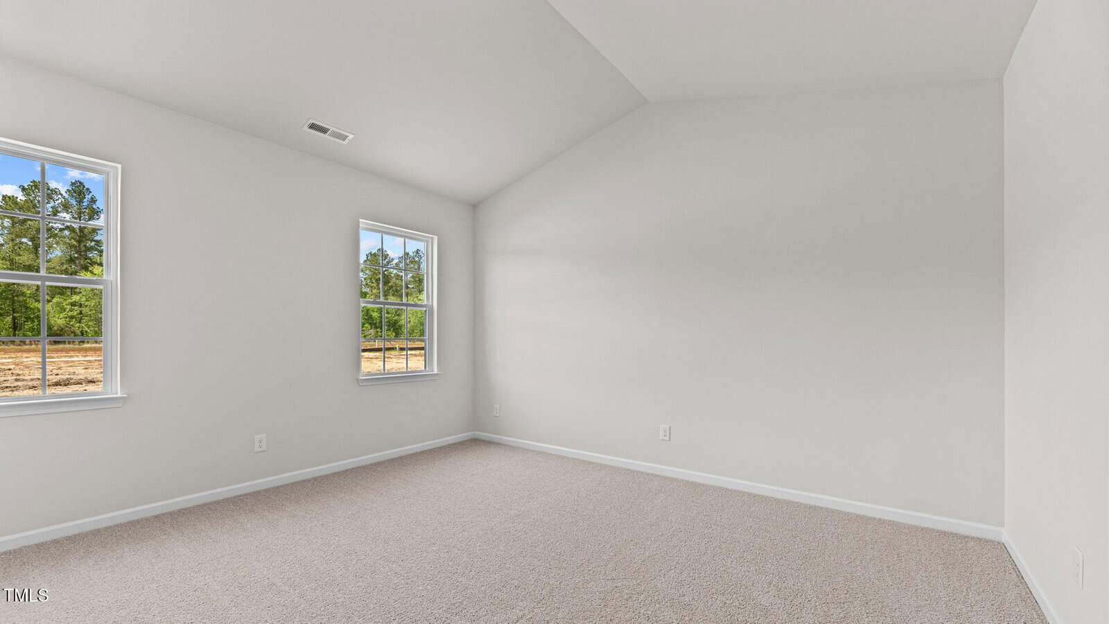 An empty room with light beige carpet, white walls, a vaulted ceiling, and two windows showing trees and a field outside. The space is well-lit with natural light.