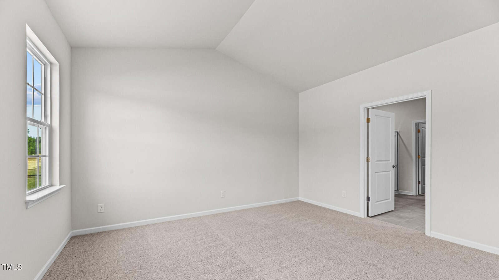 An empty room with beige carpet, white walls, a large window on the left letting in natural light, and an open white door leading to a tiled hallway or another room. The ceiling is vaulted.