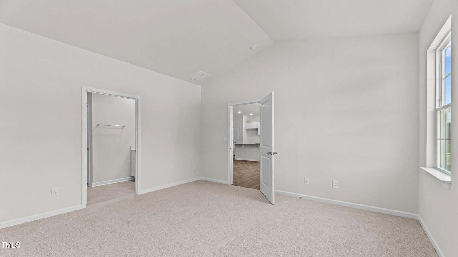 Bright, empty bedroom with beige carpet and white walls, featuring two open doors—one to a closet, another to a hallway with a view of a kitchen. Sunlight streams in through a window on the right.