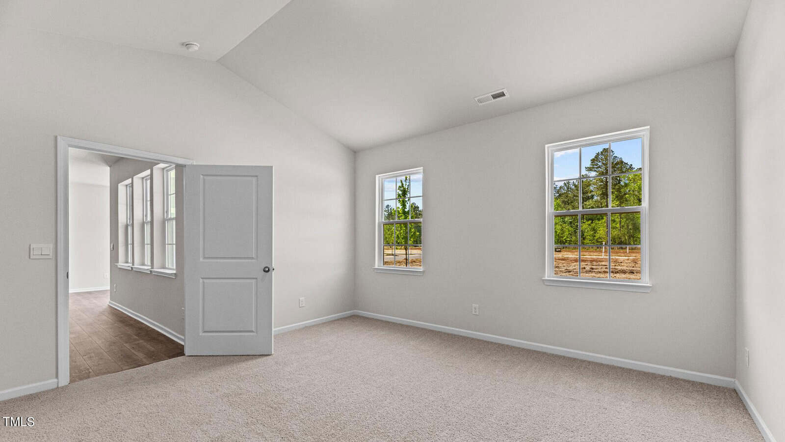 Bright, empty room with beige carpet, white walls, and two large windows showing trees outside. An open door leads to a hallway with wood flooring and additional windows. The ceiling is vaulted.