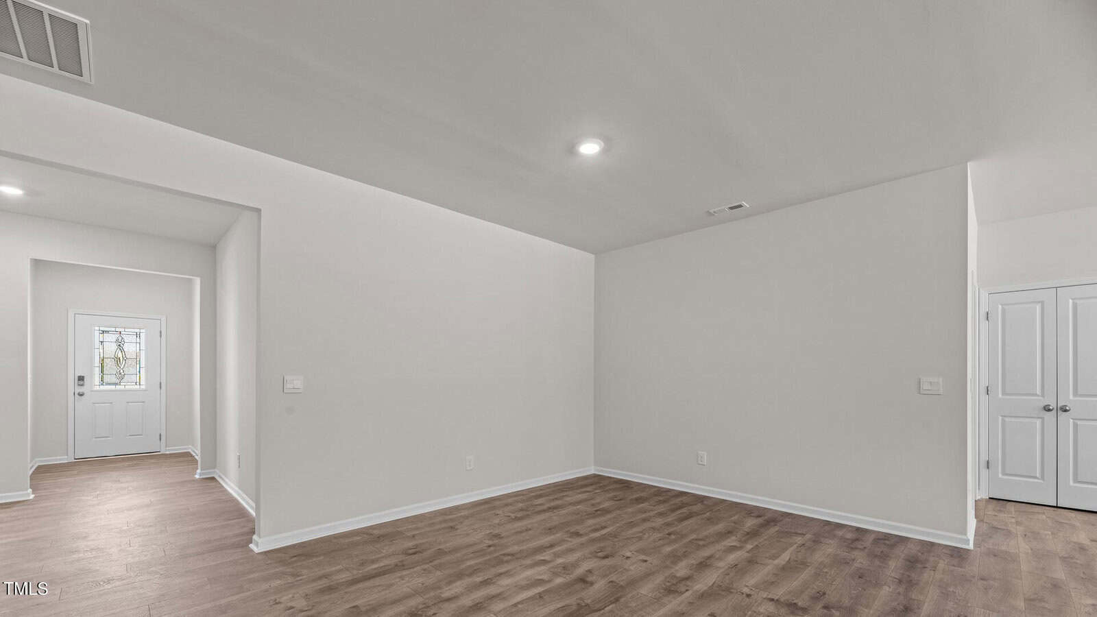 A room with light-colored walls and wood flooring, featuring recessed ceiling lights. A hallway leads to a white door with a window, and two white double doors are visible on the right. The space is empty.