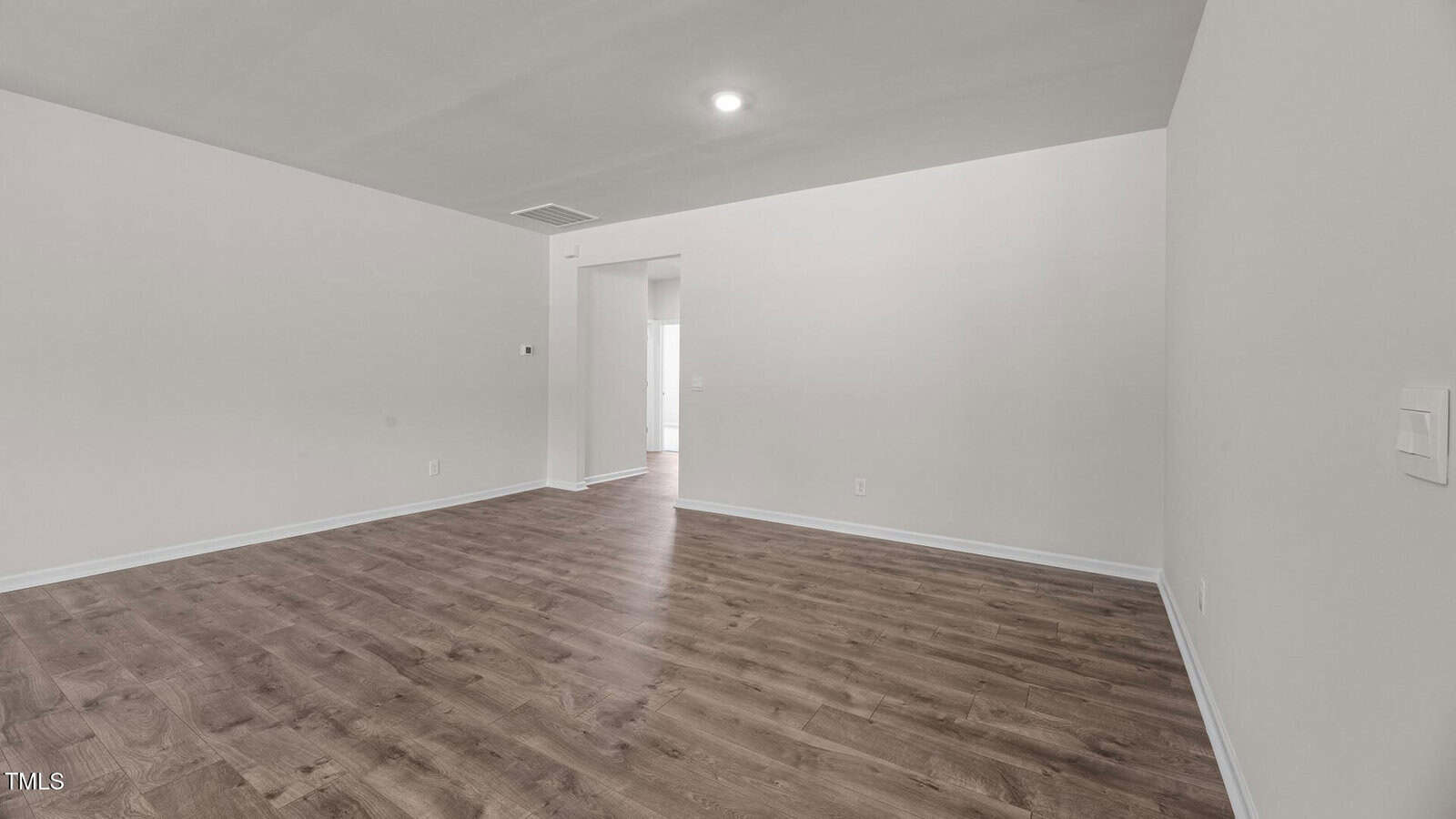 A vacant room with light gray walls, a white ceiling, recessed lighting, and wood-style flooring. An open doorway leads to another area with visible natural light. There are power outlets and a light switch on the walls.