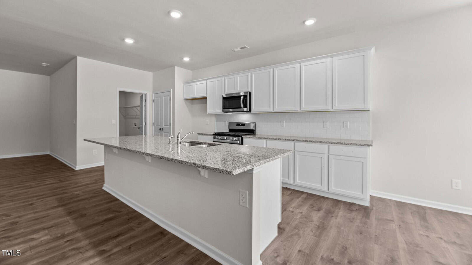A modern kitchen with white cabinets, granite countertops, stainless steel appliances, a center island with a sink, tile backsplash, and wood-style flooring under recessed ceiling lights.