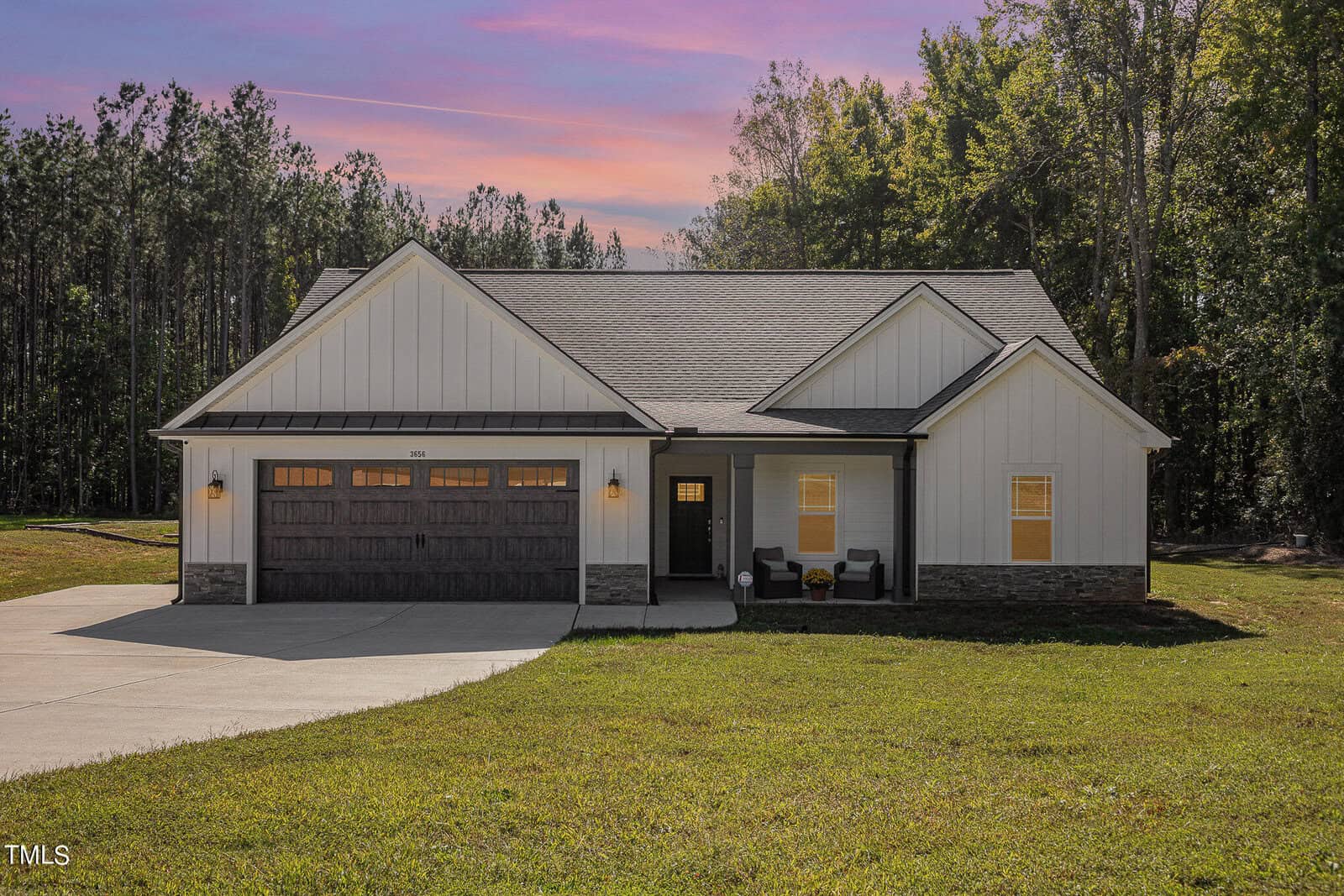 A single-story house on 3656 Bruce Garner Road features white siding and a wooden garage door, beautifully captured at sunset. Located in Wake Forest, the home boasts a small front porch with chairs, surrounded by trees and a grassy lawn.