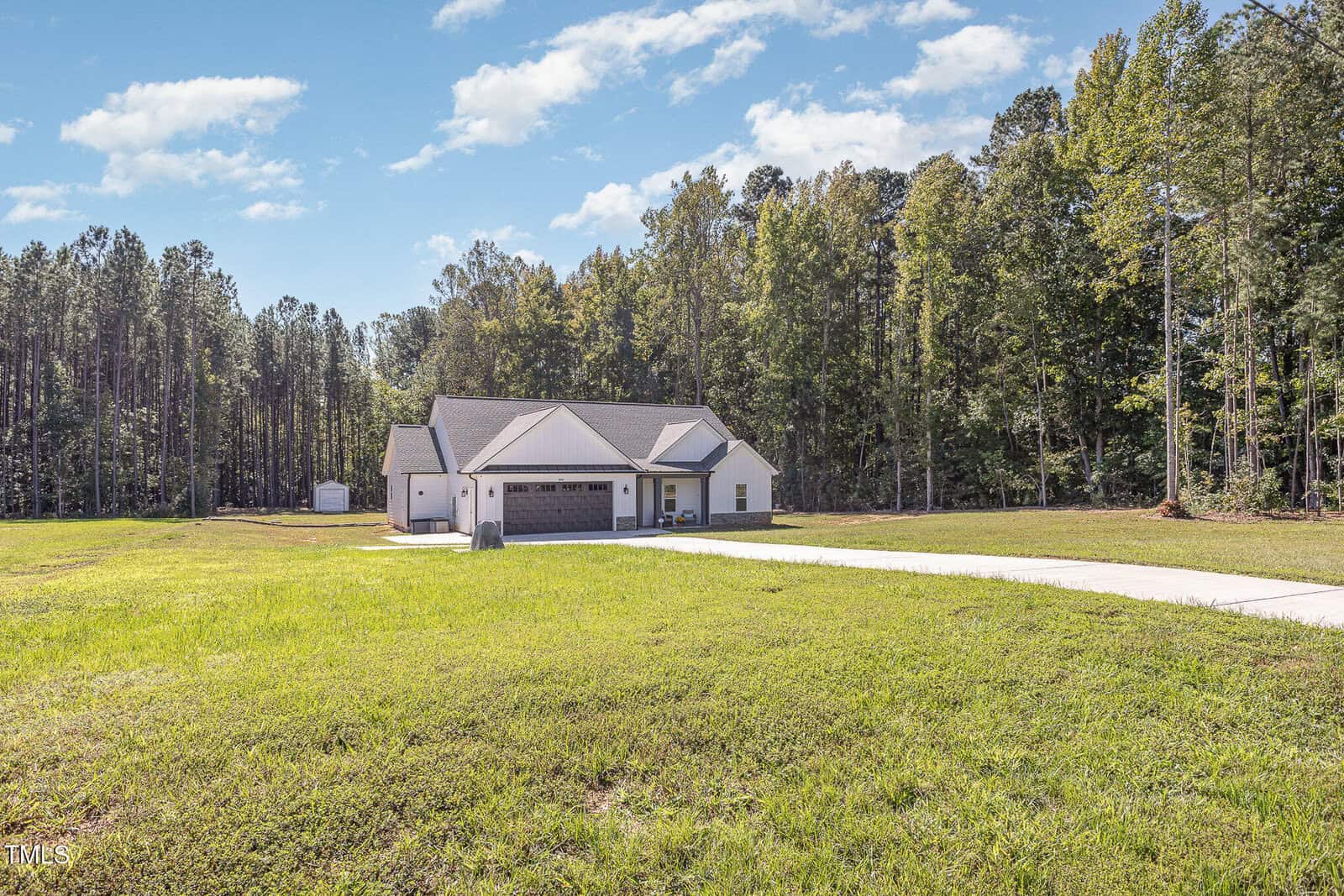 A single-story white house with a gray roof is set against a backdrop of tall trees. It features a two-car garage and a large, neatly mowed lawn. A concrete driveway leads to the garage, and the sky is clear with a few clouds.
