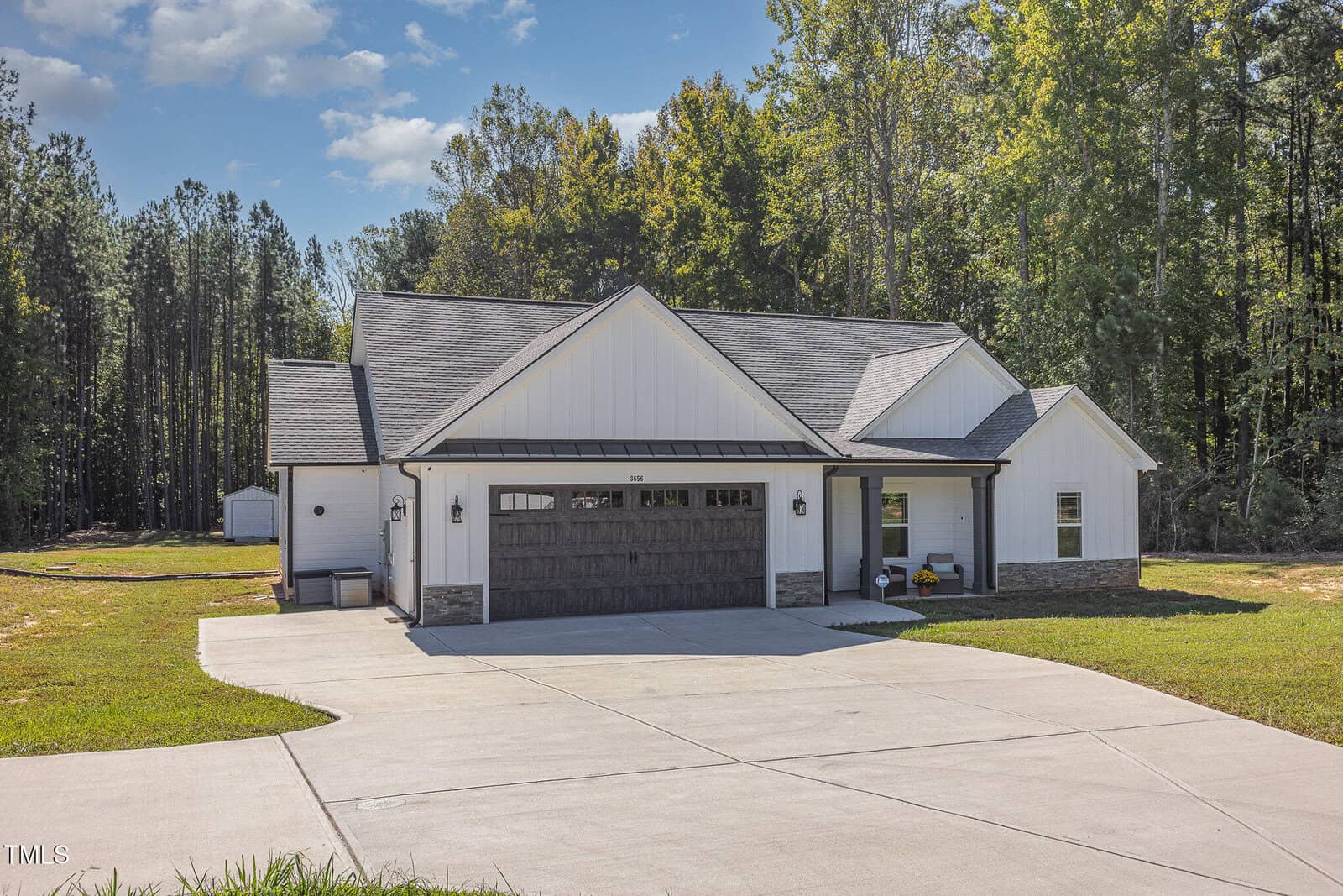 A modern, single-story house with a gray roof and white siding is set against a backdrop of tall trees. It features a two-car garage with wooden doors and a wide driveway. The sky is partly cloudy.