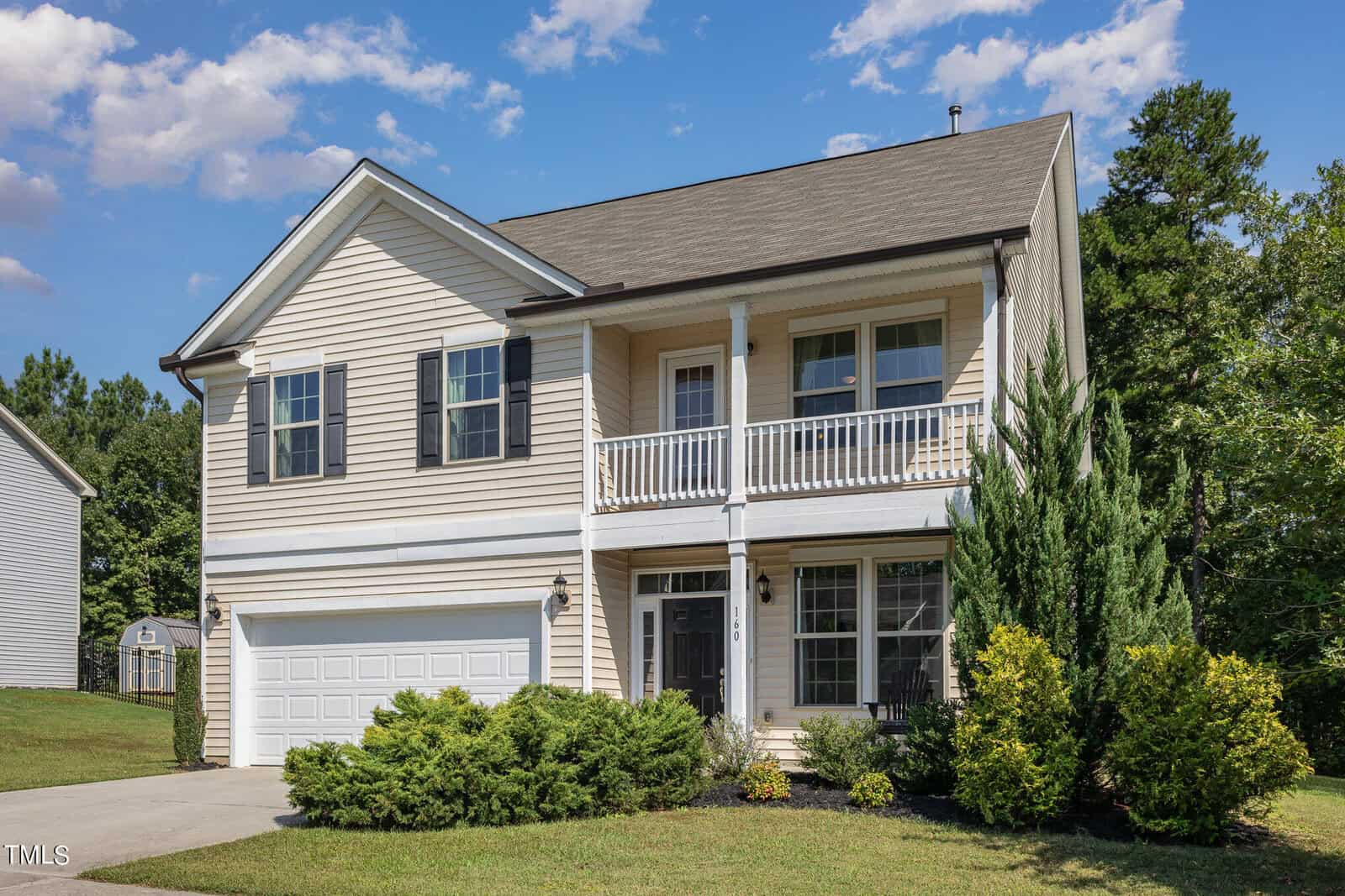 A two-story beige house with a double garage, black shutters, a second-floor balcony, and landscaped bushes in front. The house is set on a grassy lawn with trees and a blue sky with some clouds in the background.