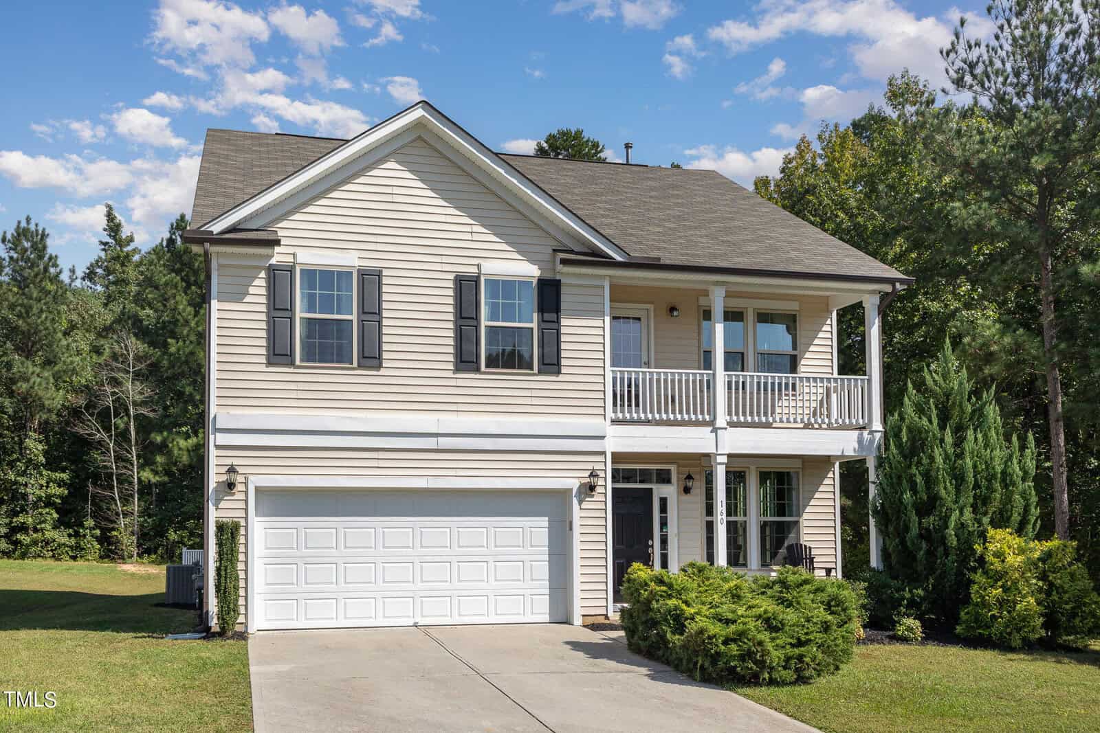 Two-story beige house with black shutters, a double garage, and a covered balcony over the front porch. The house is surrounded by grass, bushes, and trees, with a concrete driveway leading to the garage.