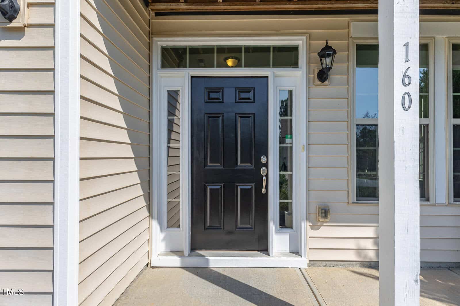 Front entryway of a house featuring a black door with side windows, beige siding, a white door frame, a black wall-mounted light fixture, house number 160, and a covered porch area with concrete flooring.