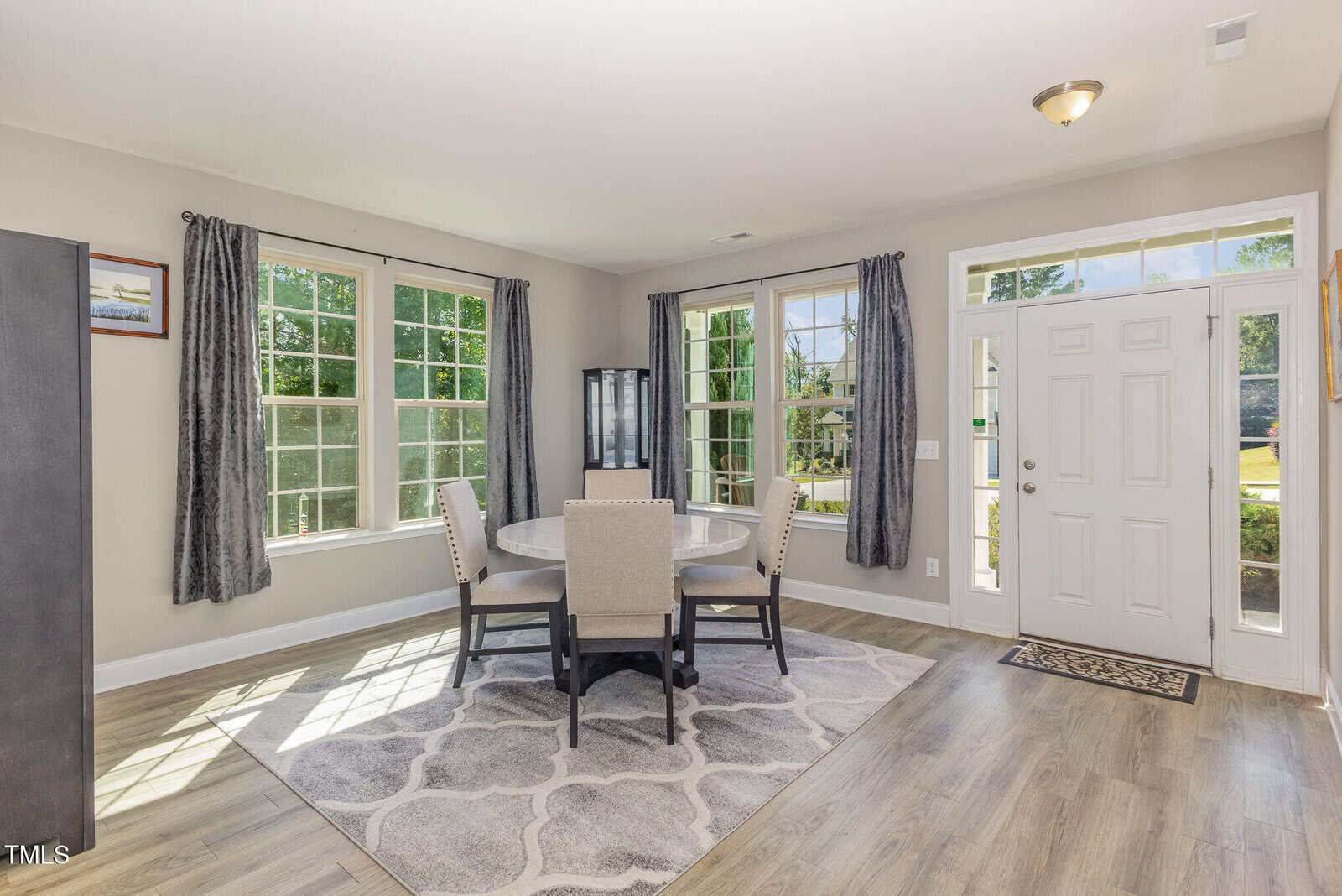 A bright room with three large windows, a round dining table with four upholstered chairs, gray curtains, a patterned rug, wood-look flooring, and a white front door with sidelights and a transom window.