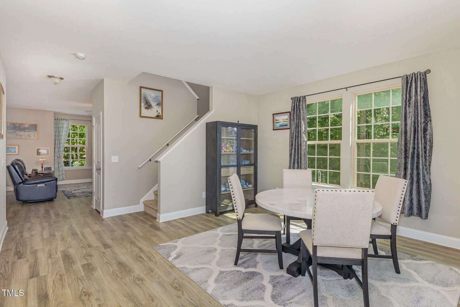 A dining area with a round table and four upholstered chairs sits next to large windows with gray curtains. Nearby is a glass display cabinet, and a staircase leads to the upper floor. Hardwood floors and neutral walls complete the space.