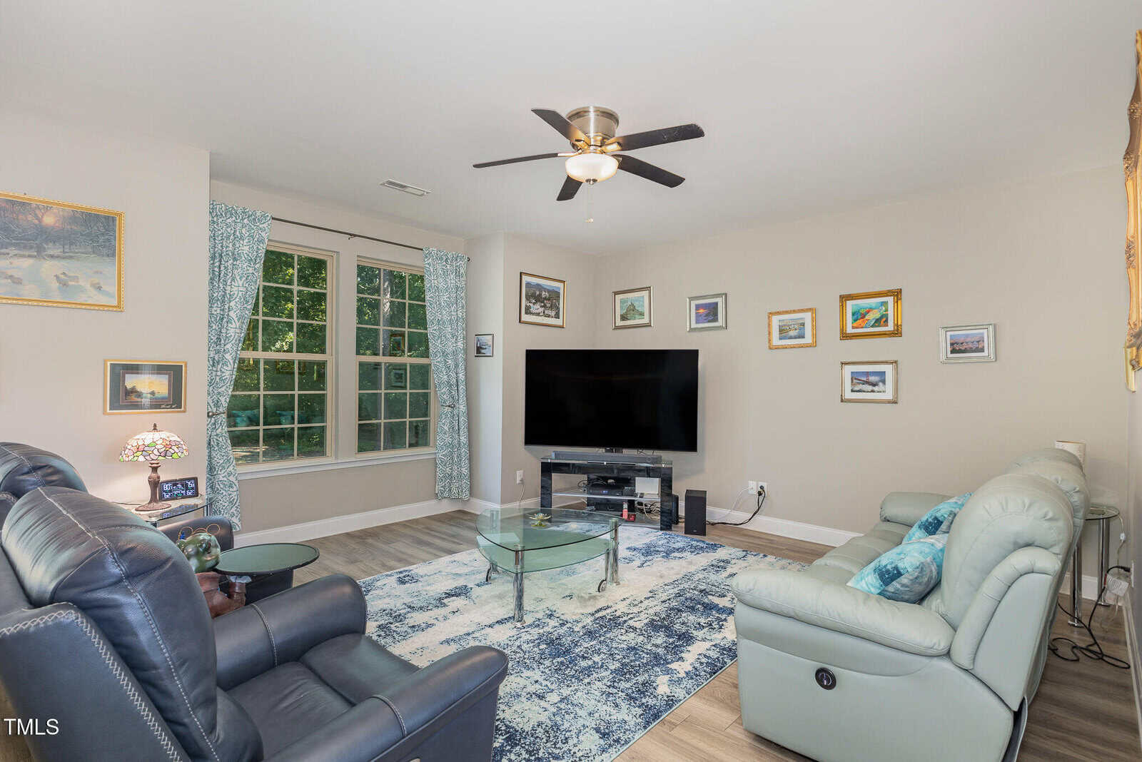 A living room with light walls, a large window with blue patterned curtains, a ceiling fan, a gray sectional sofa, a glass coffee table, and a flat-screen TV on a stand. Framed pictures decorate the walls.