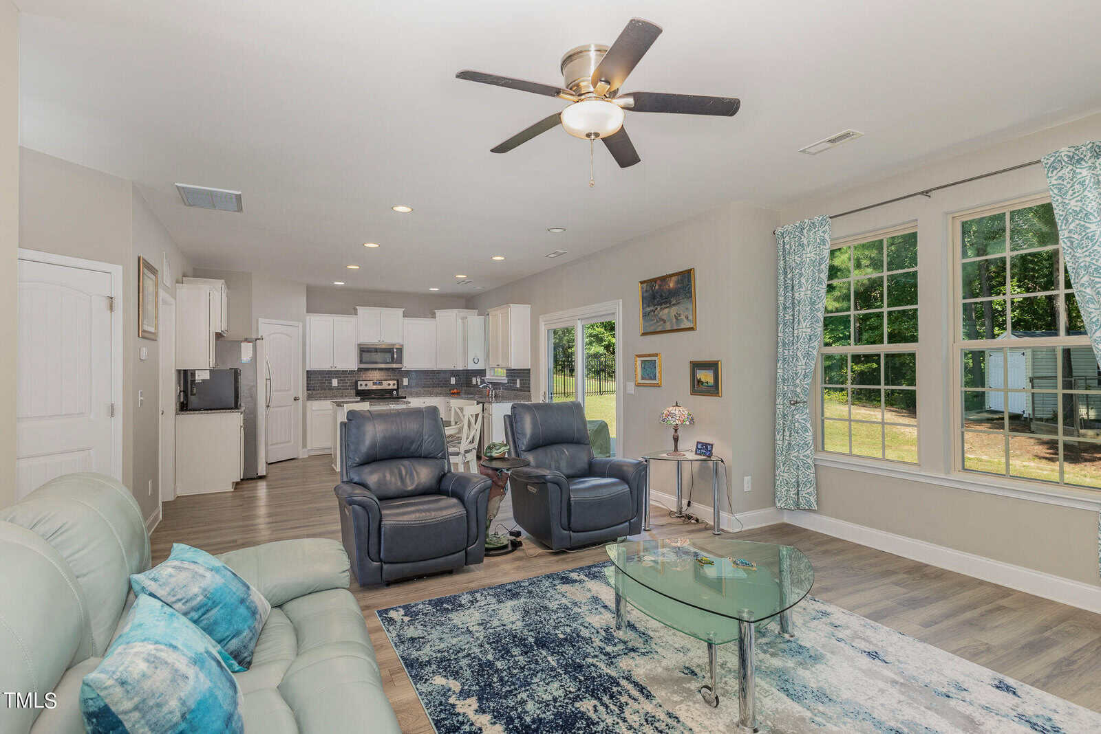 A bright, modern living room with light walls, a ceiling fan, two blue recliners, a light sofa, a glass coffee table, and an open kitchen in the background. Large windows let in natural light.