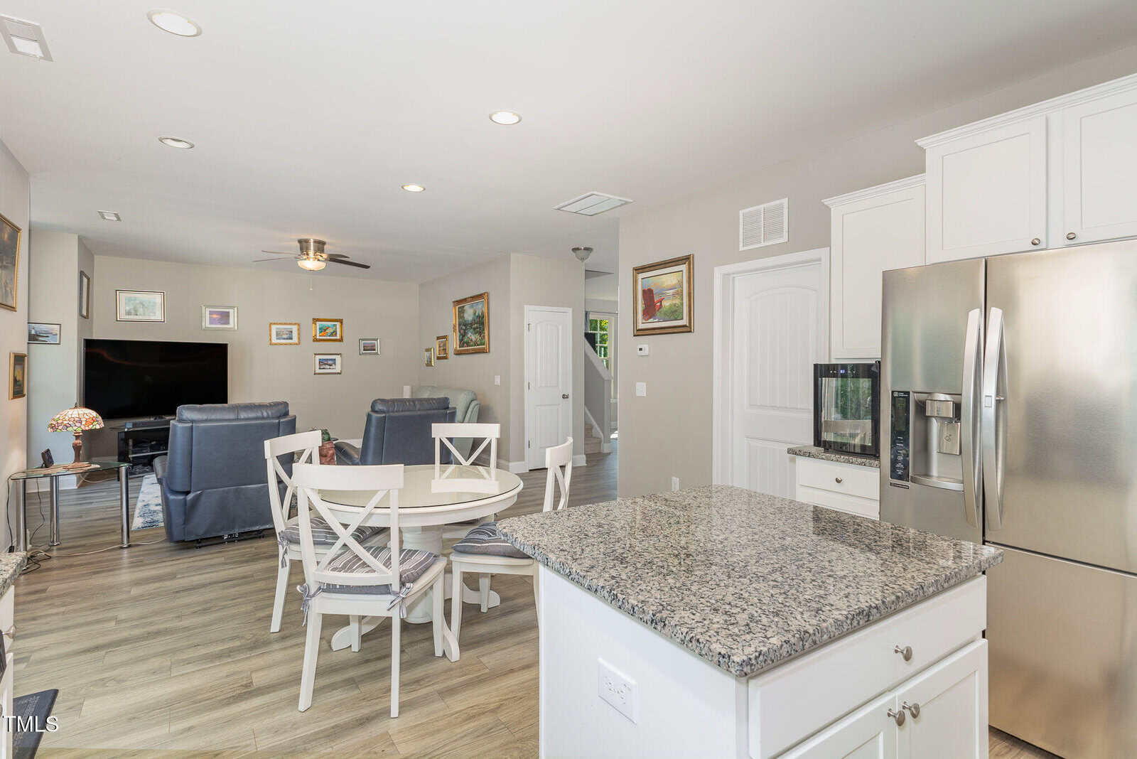Open-concept kitchen and living area with granite countertops, a round dining table with four chairs, stainless steel refrigerator, light gray flooring, and various framed pictures on the walls.