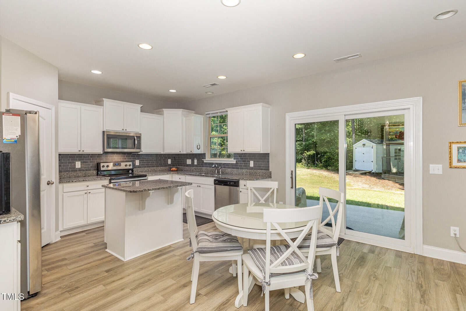 Modern kitchen with white cabinets, stainless steel appliances, a small island, and a round dining table with four chairs. Glass sliding doors open to a backyard with a shed visible outside. Light wood flooring throughout.