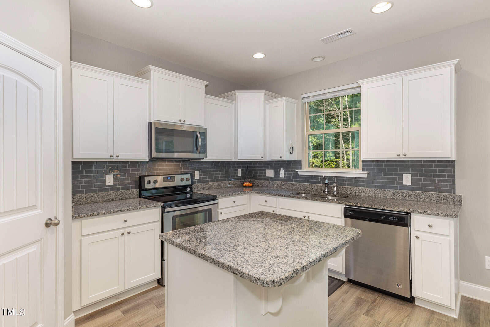 Modern kitchen with white cabinets, stainless steel appliances, gray tile backsplash, granite countertops, and a center island. A window above the sink lets in natural light, and the floor is a light wood.
