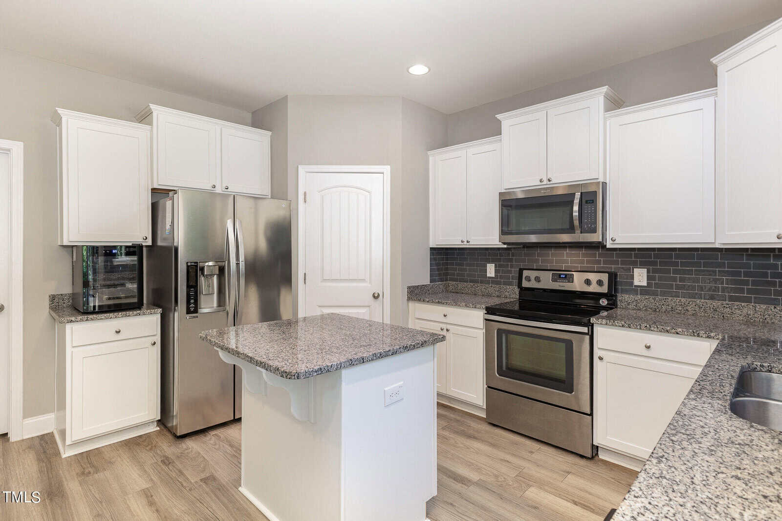 Modern kitchen with white cabinets, stainless steel appliances, granite countertops, a small island, gray tile backsplash, and light wood flooring. A closed white door is centered in the background.