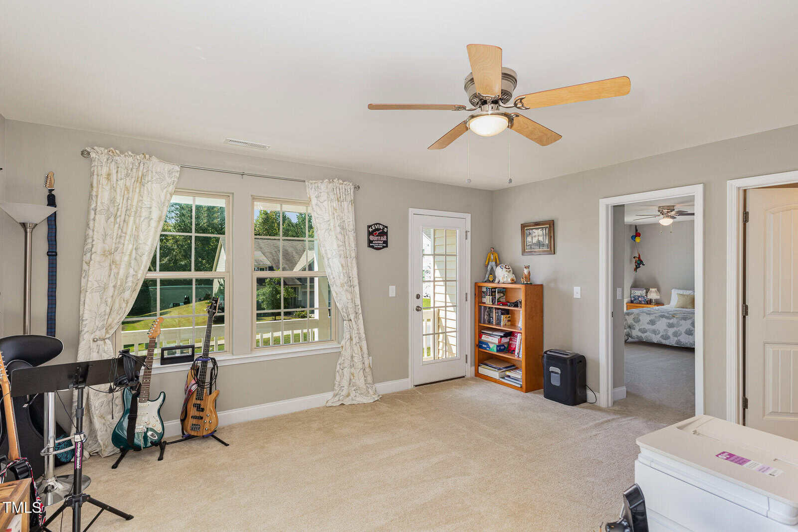 A bright, carpeted room with a ceiling fan, two windows with curtains, a glass door, guitars and a stand, a bookshelf, and a doorway leading to another room with a bed.