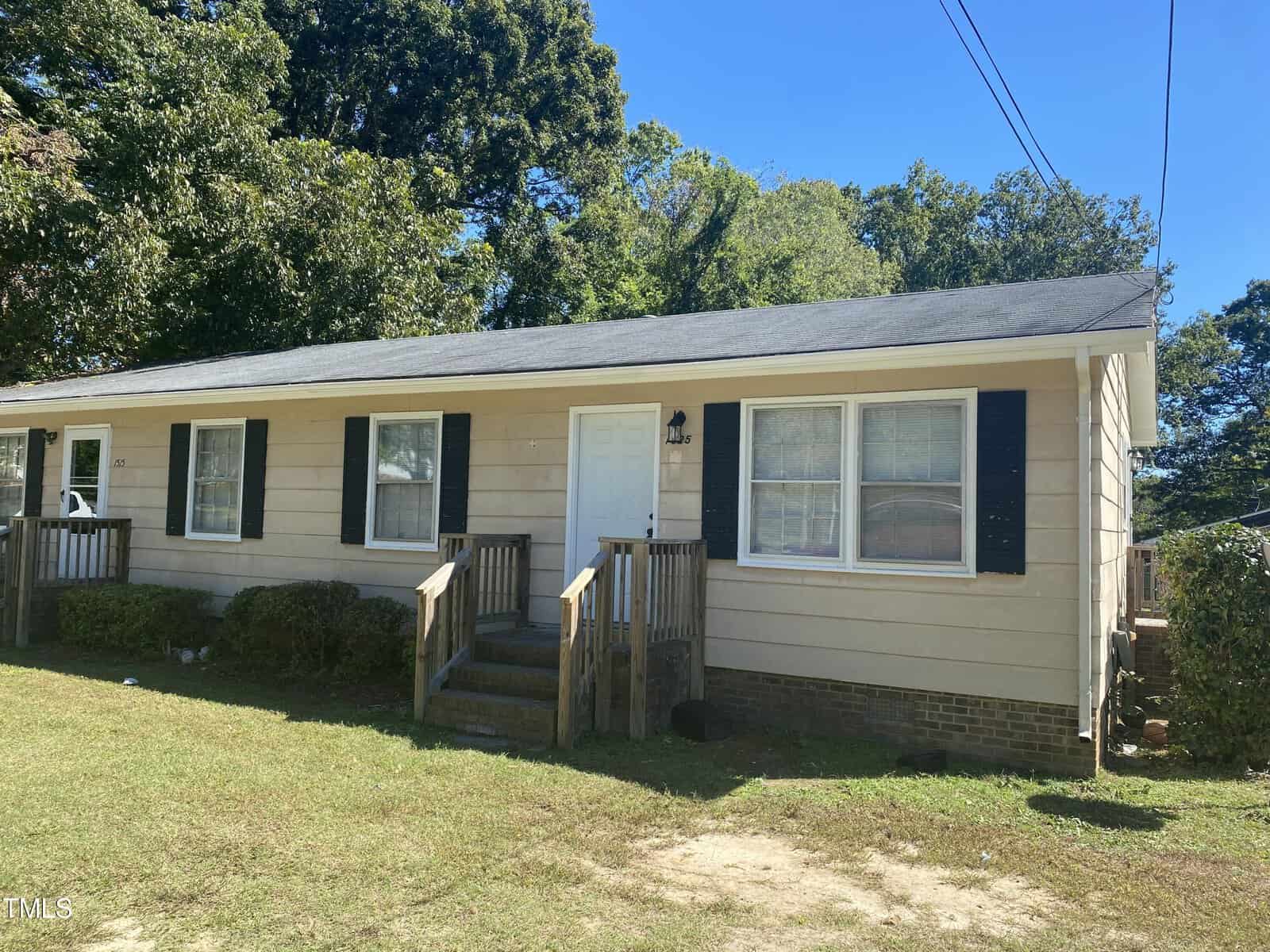 The one-story beige house at 1515 Sunset Avenue in Henderson boasts dark shutters and a shingled roof. It features a small wooden porch with steps leading to the front door, surrounded by grass and trees under a clear blue sky.