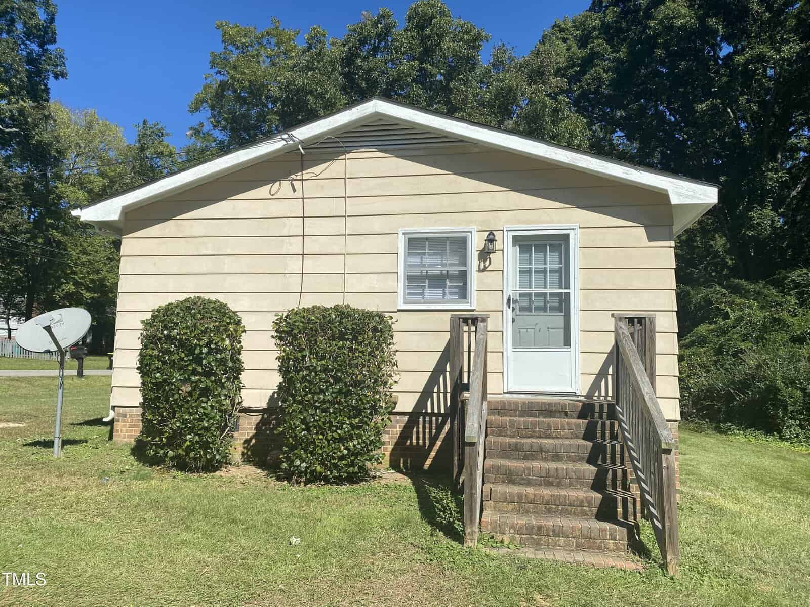 Small beige house with white trim, one front window, and a white door. Steps lead to the entrance, flanked by two large bushes. A satellite dish is on the left lawn. Tall trees are in the background under a clear blue sky.