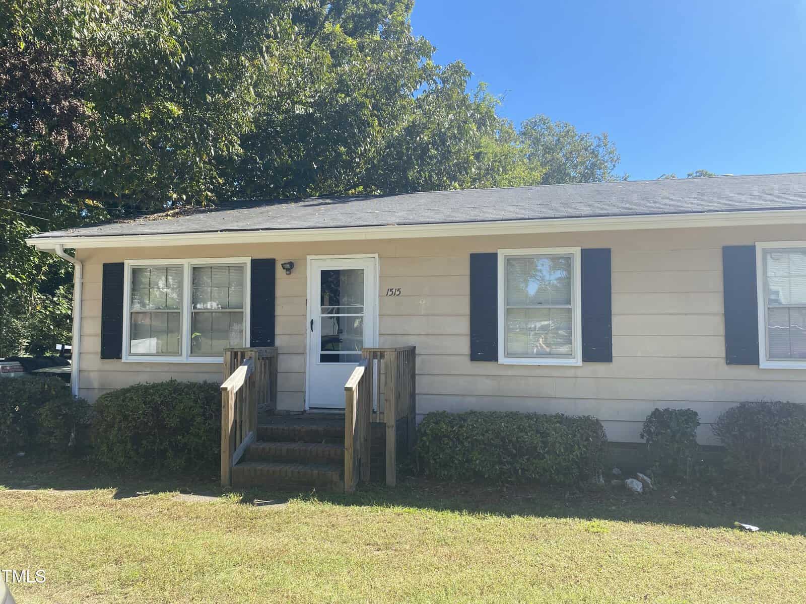 A small beige house with dark shutters and a shingled roof. It has a front porch with wooden steps leading up to a glass front door. The house is surrounded by neatly trimmed bushes and trees in the background under a clear blue sky.