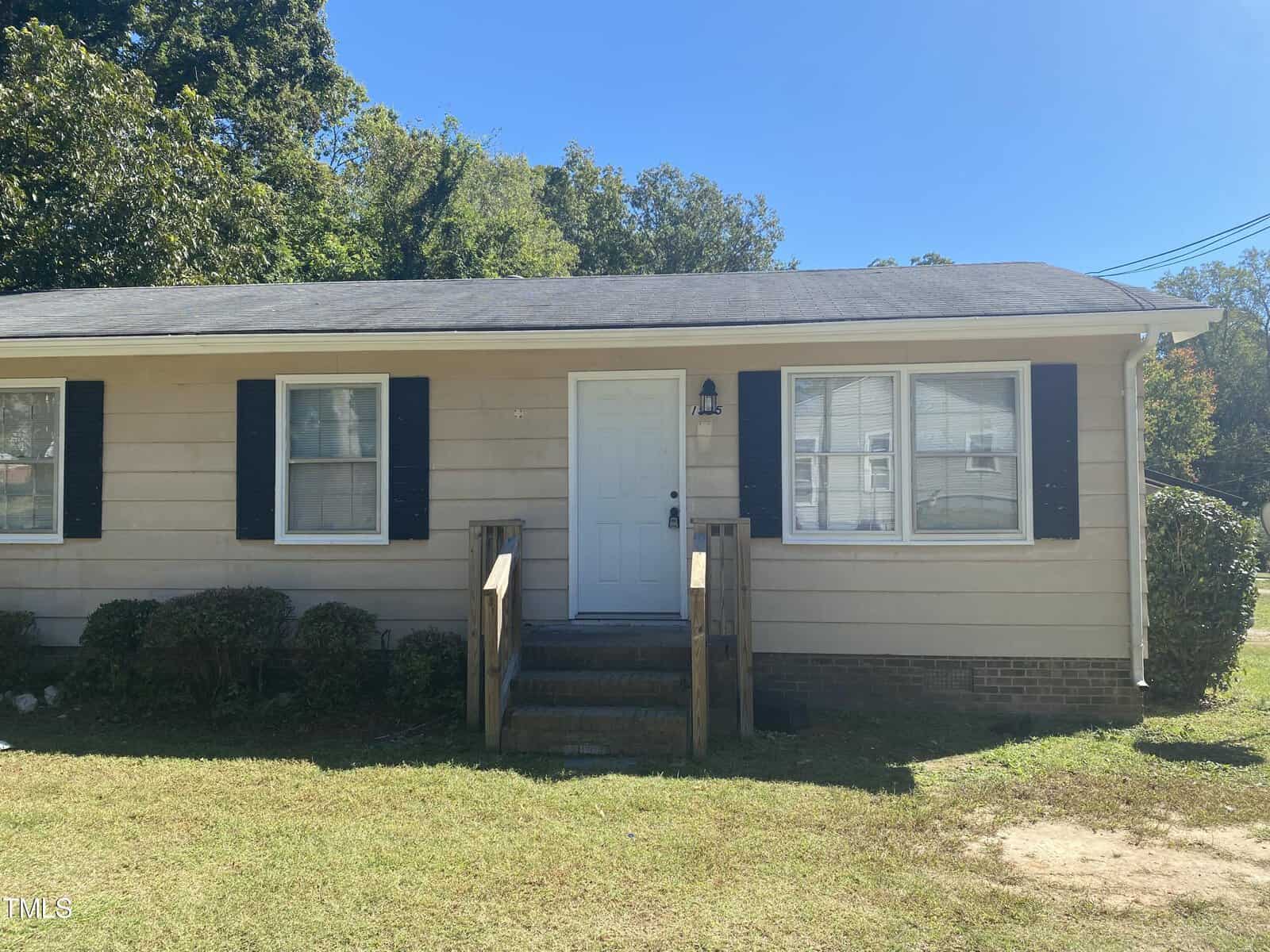A single-story beige house with a dark roof, white door, and black shutters. It has a small porch with wooden steps and a grassy front yard. Trees are in the background under a clear blue sky.