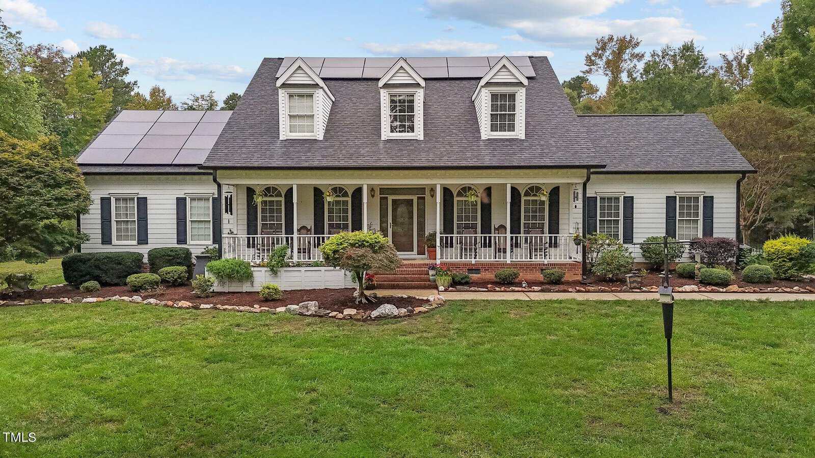 A large white house at 4077 Range Road boasts a symmetrical design, featuring a covered front porch and multiple dormer windows. The solar-paneled roof complements its eco-friendly appeal. Nestled behind a well-maintained lawn and garden, it's surrounded by trees under a partly cloudy sky.