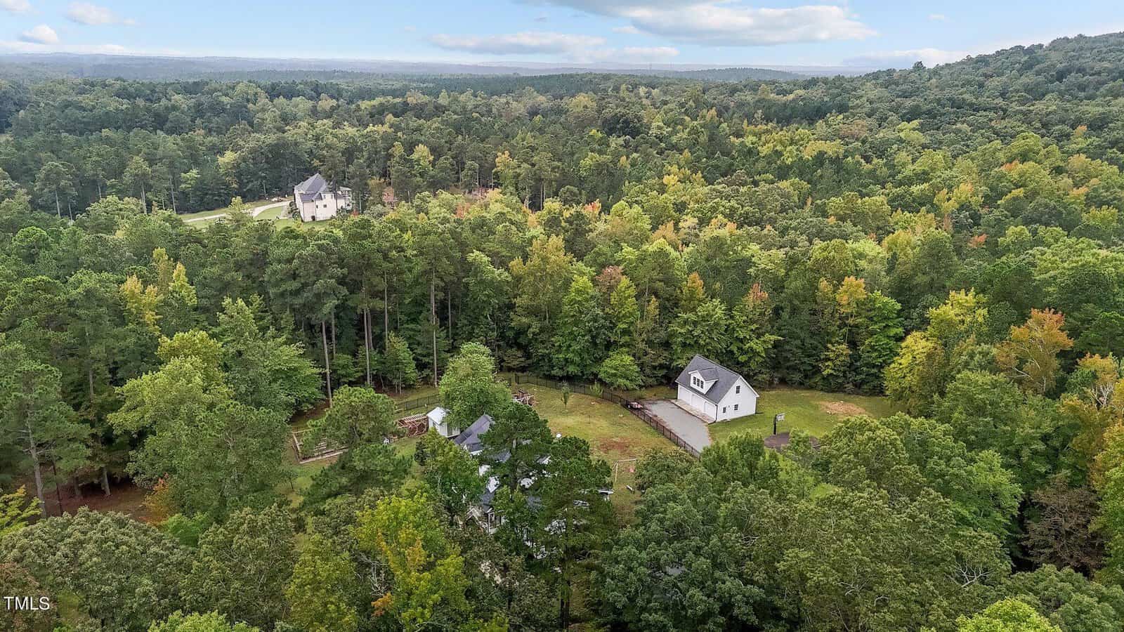 Aerial view of a forested landscape with several houses scattered among the trees. A clear sky with some clouds is visible above. The area appears lush and green, suggesting a rural or suburban setting.