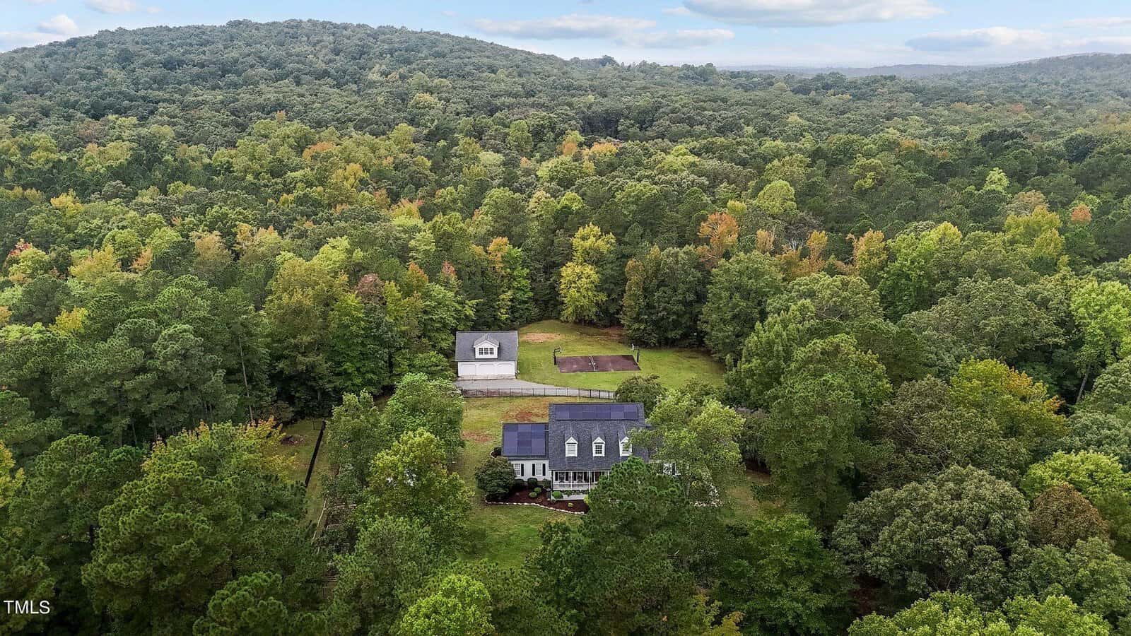 Aerial view of a large house surrounded by dense green forest. The property includes a smaller building and an open field with visible pathways. Lush hills rise in the background under a cloudy sky.