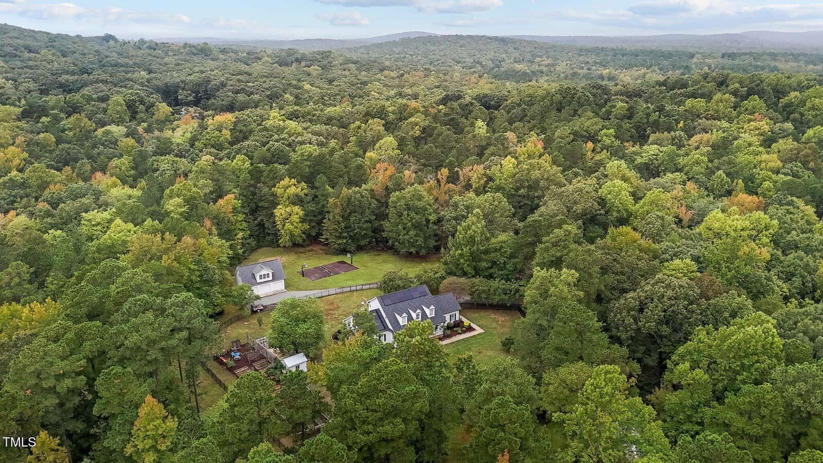 Aerial view of a house surrounded by dense green forest. The property includes a separate garage, a garden, and appears to be isolated within a vast area of trees. Mountains are visible in the distance under a partly cloudy sky.