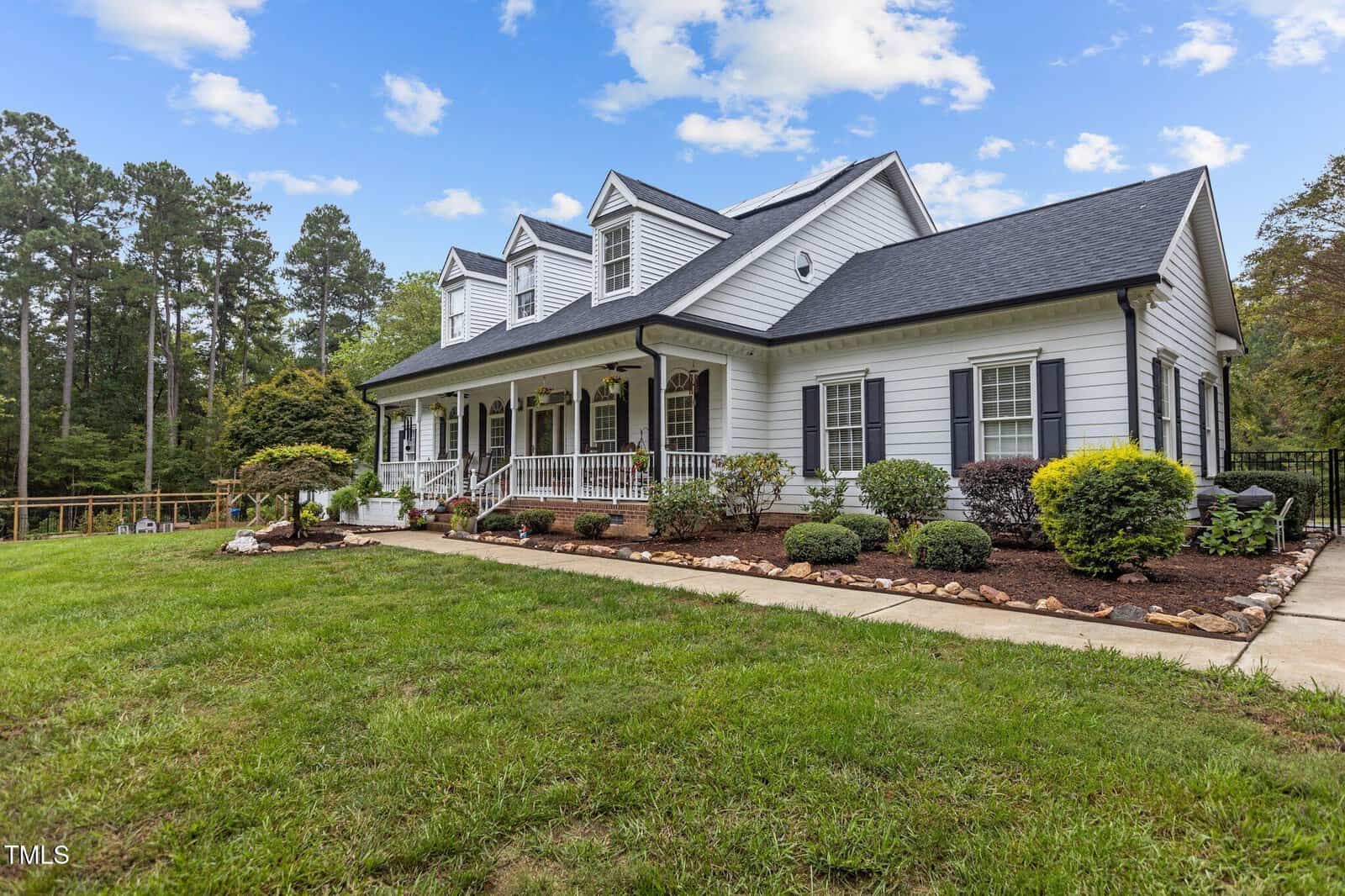 A two-story white house with black shutters and a dark roof sits in a landscaped yard. The home features a covered porch with several windows and dormers. The lawn is well-kept, and there are shrubs and trees around the walkway.