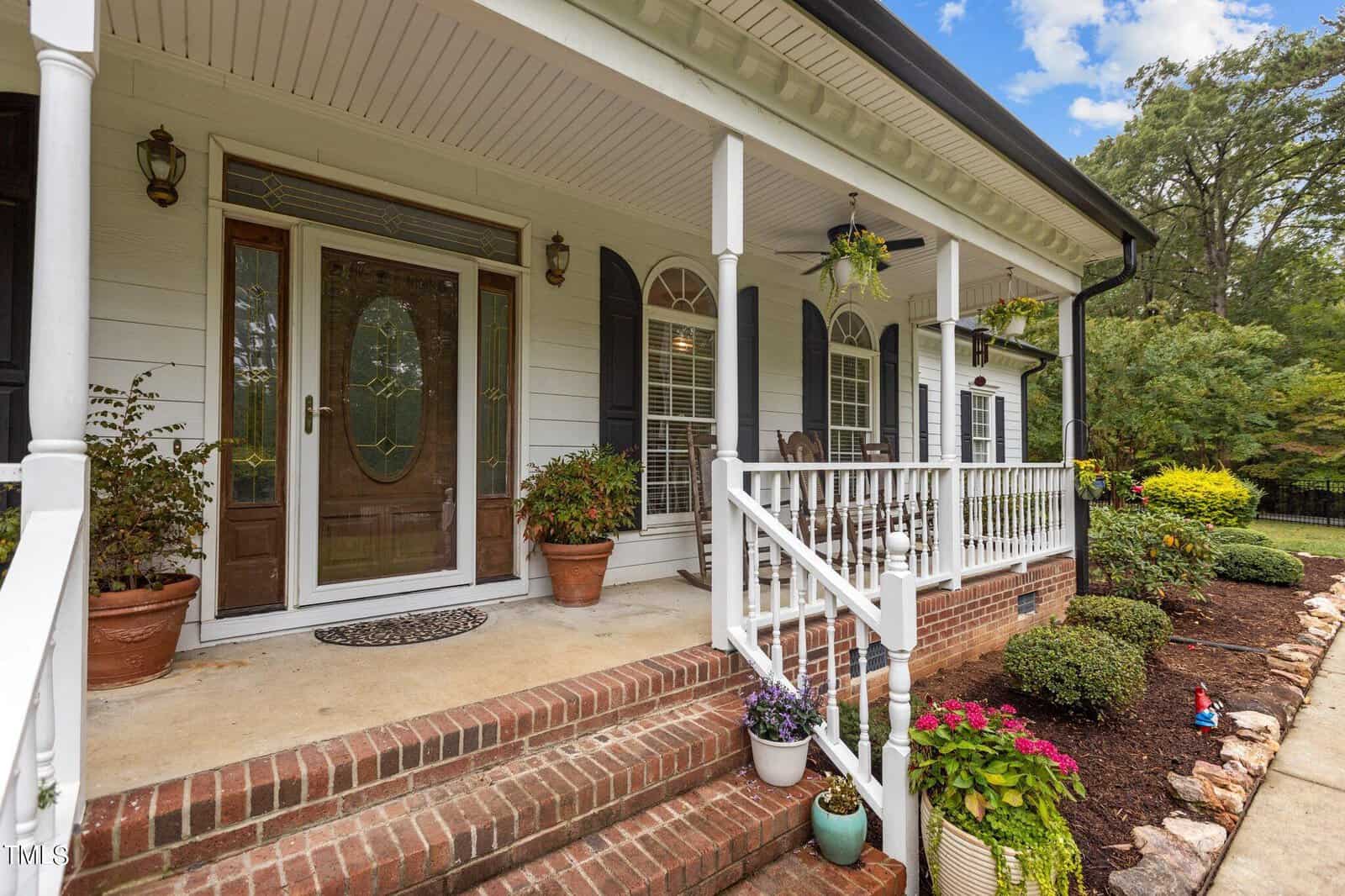 A white house features a covered front porch with brick steps and white railings. Two chairs are placed on the porch, surrounded by potted plants. The front door includes glass panels. The yard has manicured shrubs and a stone-lined garden bed.