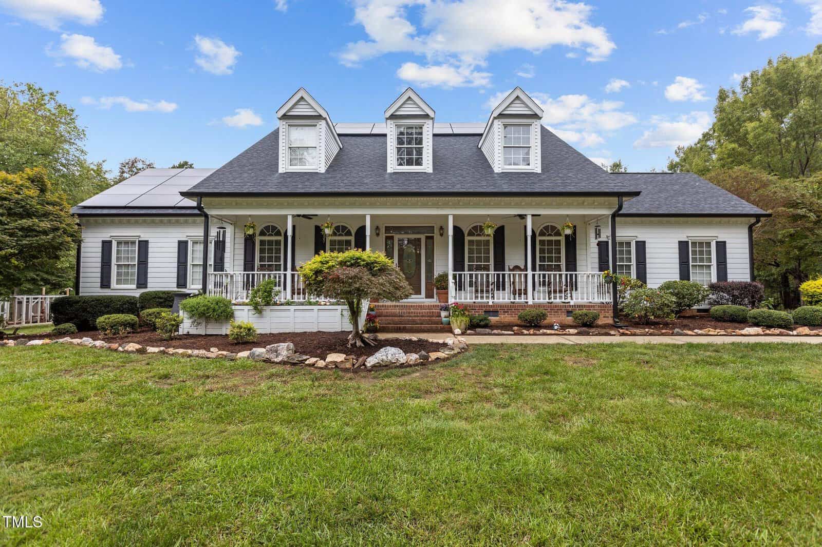 A large white house with a black roof, featuring three dormer windows, a covered front porch, and well-maintained landscaping. The lawn is spacious, and a few trees are in the front yard. The sky is partly cloudy.
