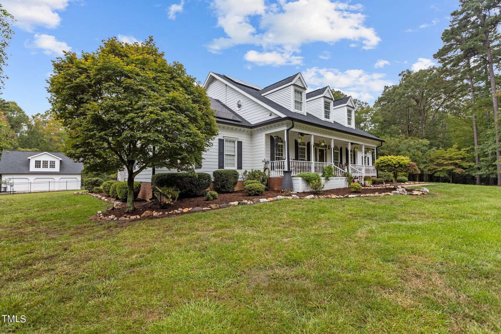 A two-story white house with a covered porch, black shutters, and a gabled roof. It's surrounded by grass and landscaped shrubs, with a detached garage visible in the background and trees on the right. The sky is partly cloudy.