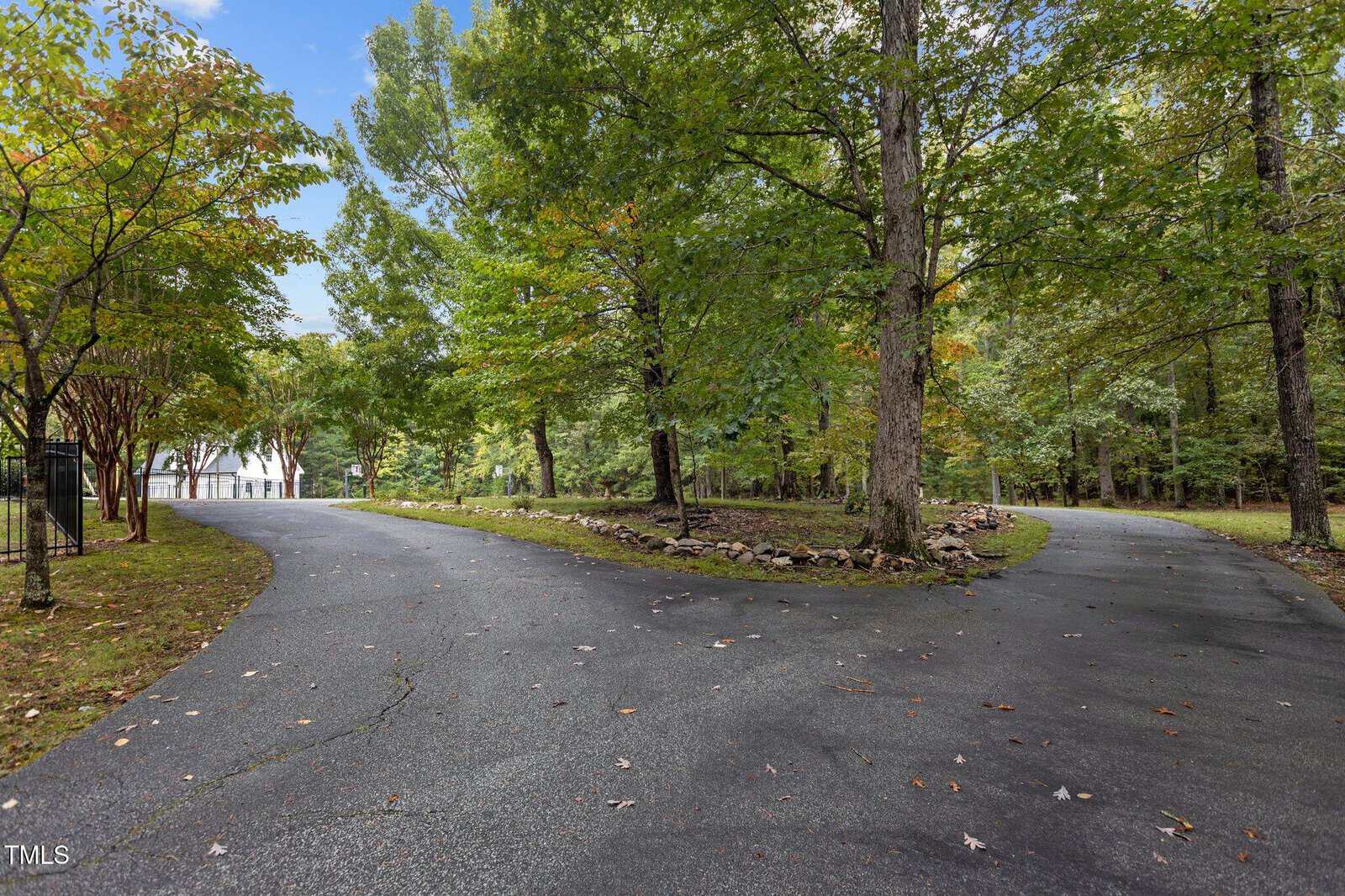 A paved driveway splits into two paths surrounded by trees with green foliage. The sky is partly cloudy, and a white building is partially visible through the trees on the left. Fallen leaves are scattered on the pavement.