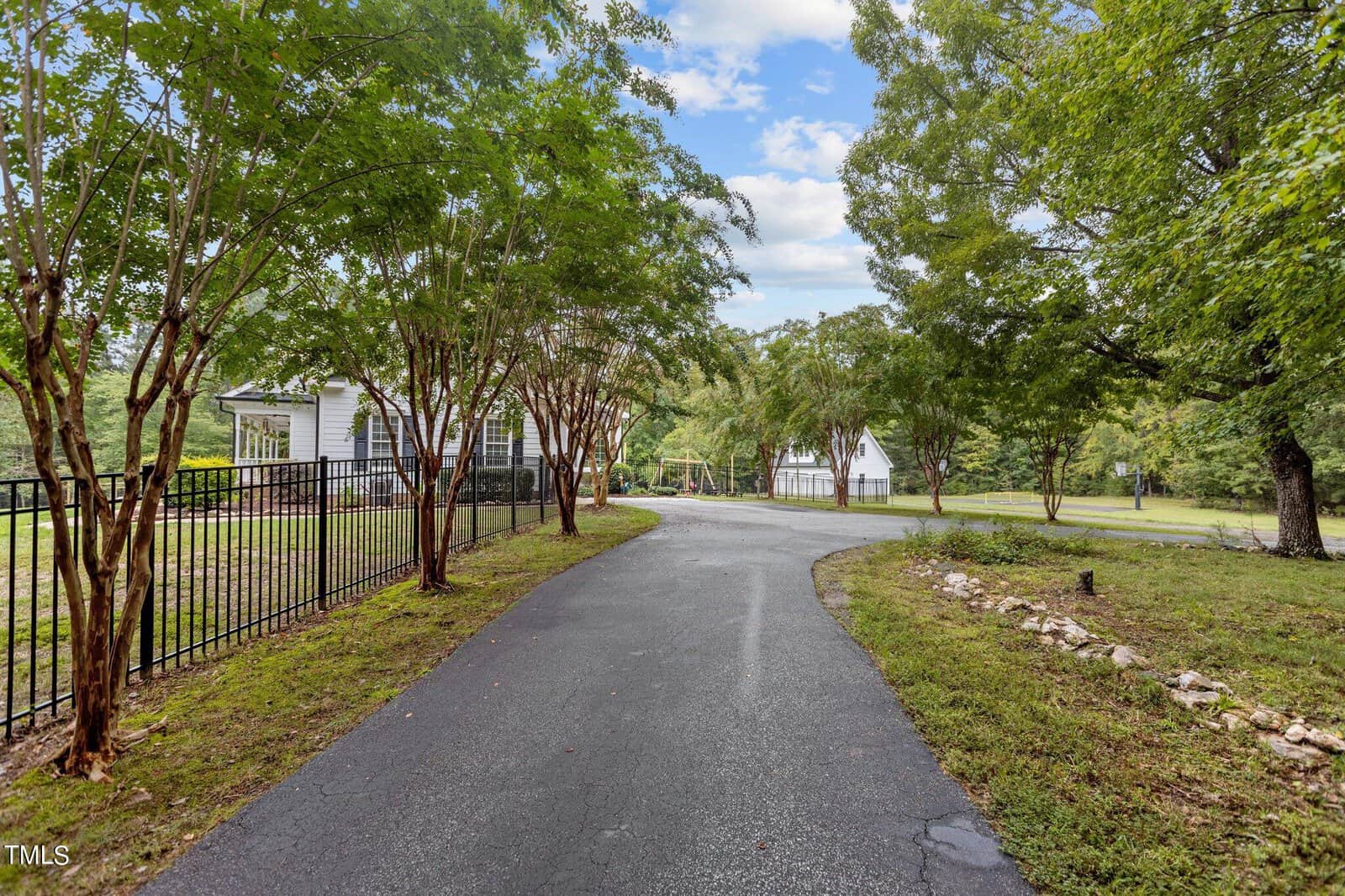 A curved asphalt driveway flanked by trees leads to houses in the background. A black metal fence borders the left side, and grass with scattered rocks lines the right. The sky is partly cloudy.