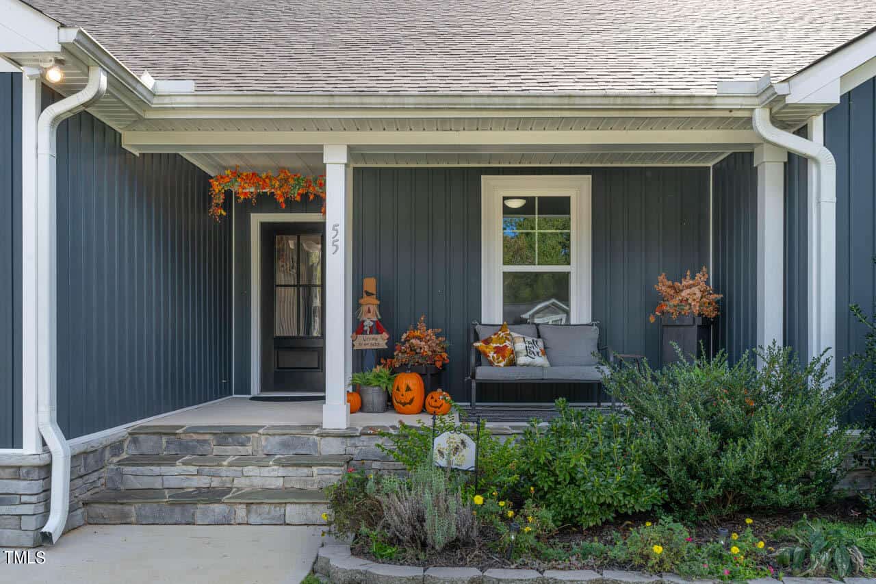 A front porch decorated for autumn with pumpkins, a scarecrow, orange foliage, and a bench with pillows. The house has dark siding, white trim, and a stone border with green shrubs in front.
