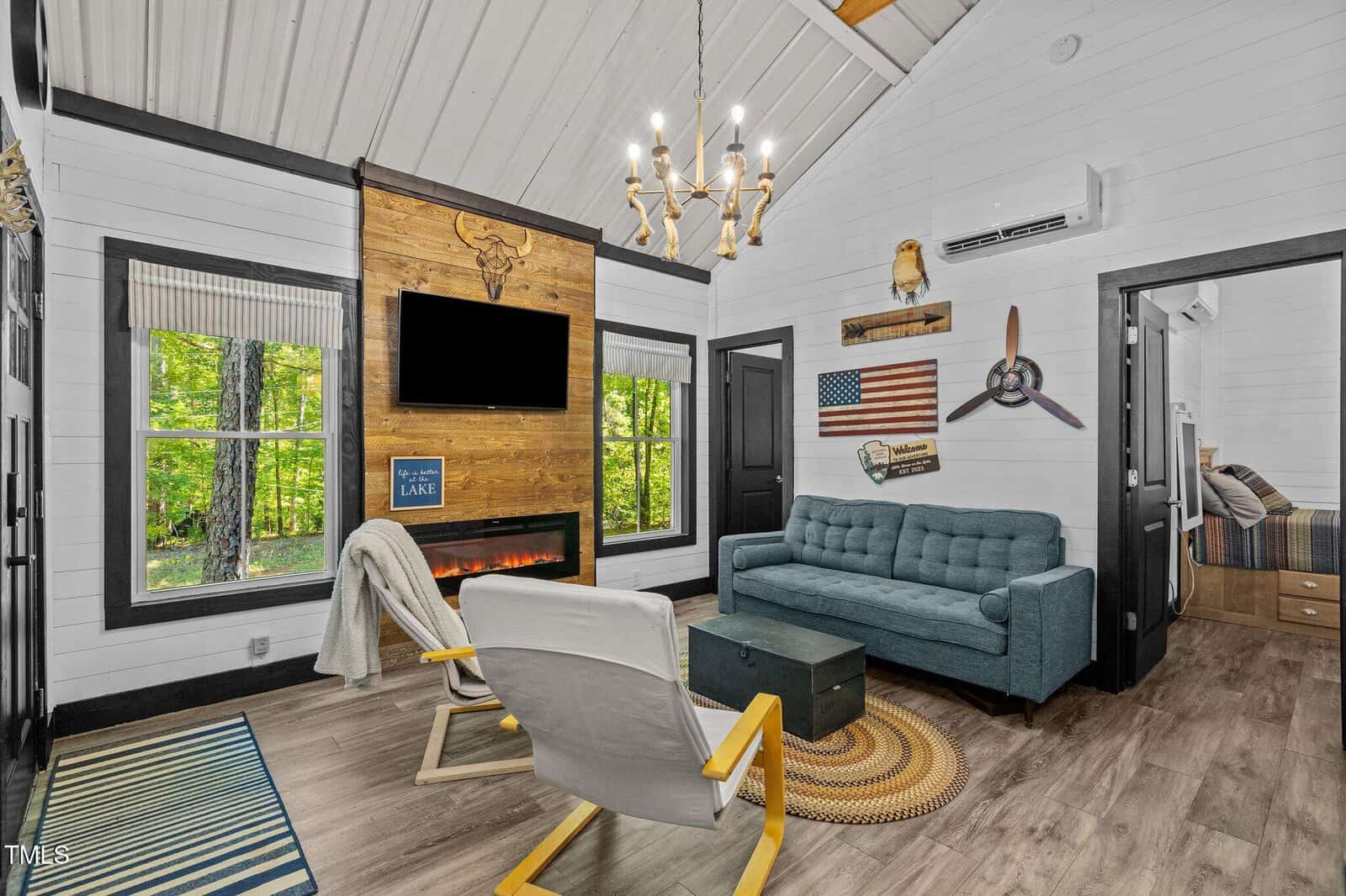 Cozy cabin living room with wood-paneled fireplace, mounted TV, blue sofa, white chair, and rustic decor. Large windows reveal green forest outside. An American flag and airplane propeller hang on the wall.