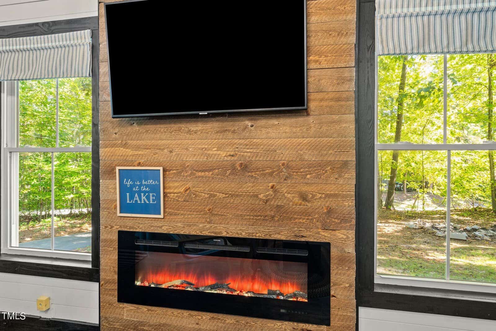 A modern wooden fireplace wall with an electric fireplace, a TV above, a blue sign saying “Life is better at the lake,” and large windows showing green trees outside.