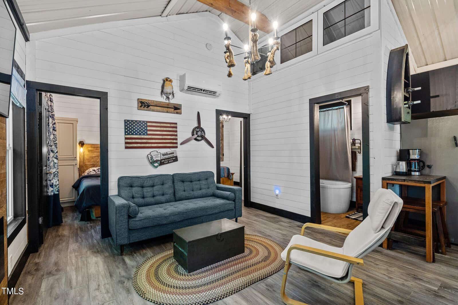 A cozy living room with a blue sofa, white armchair, round braided rug, and wall art including an American flag. The space features wood floors, white shiplap walls, and an open doorway leading to a bathroom and bedroom.