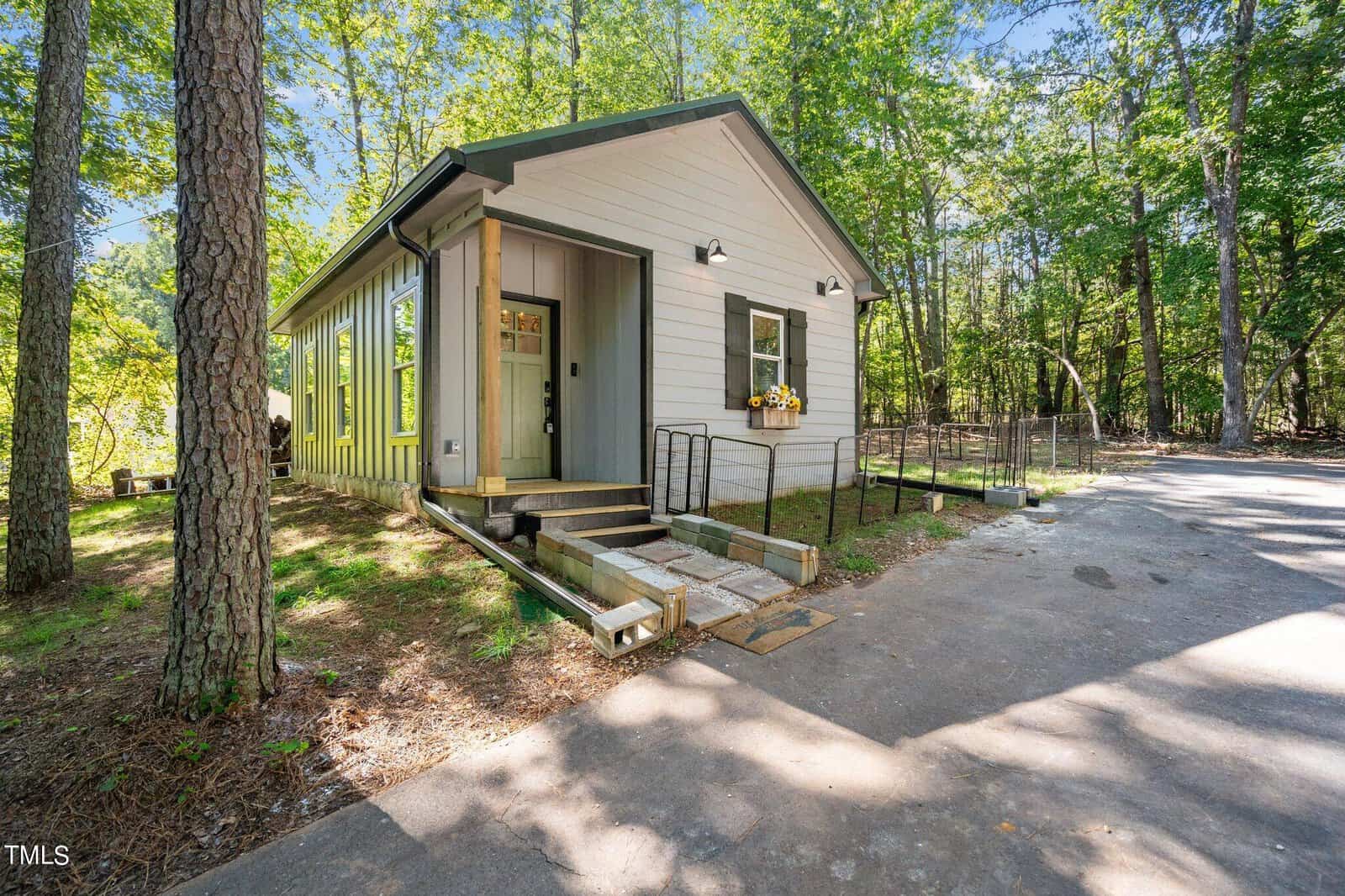 A small, light gray house with green trim and a front porch sits among tall trees. There are ramps leading to the door and a window box with yellow flowers. Sunlight filters through the surrounding forest.