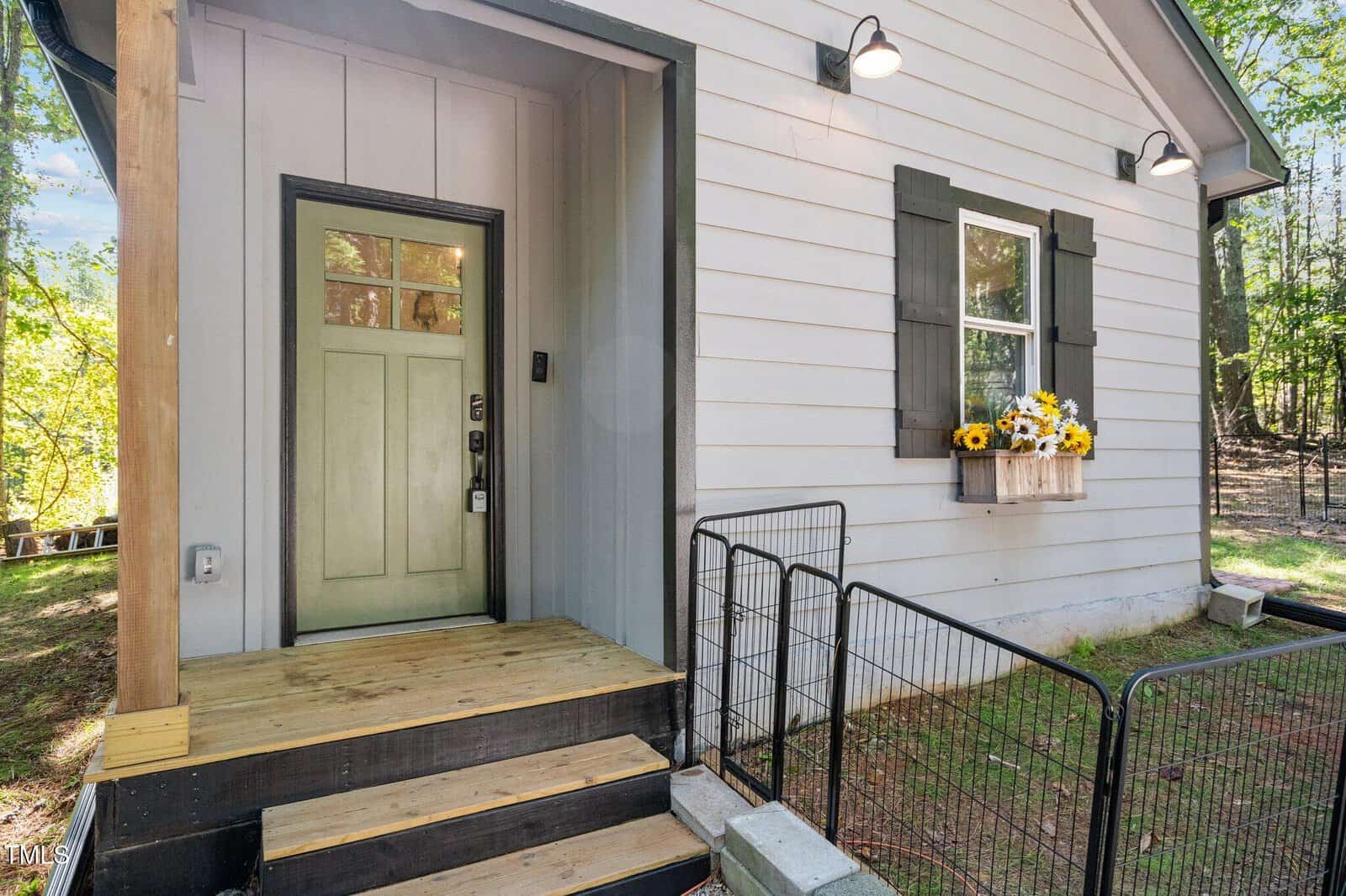 Small house entrance with a light green front door, wooden steps, and black railing; window with black shutters and a flower box holding yellow and white flowers on the right side; outdoor lights above the door and window.