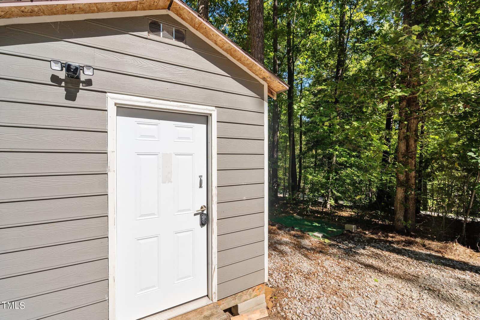 A small gray shed with a white door and security camera, set on a gravel surface near a wooded area with tall trees and sunlight filtering through the leaves.