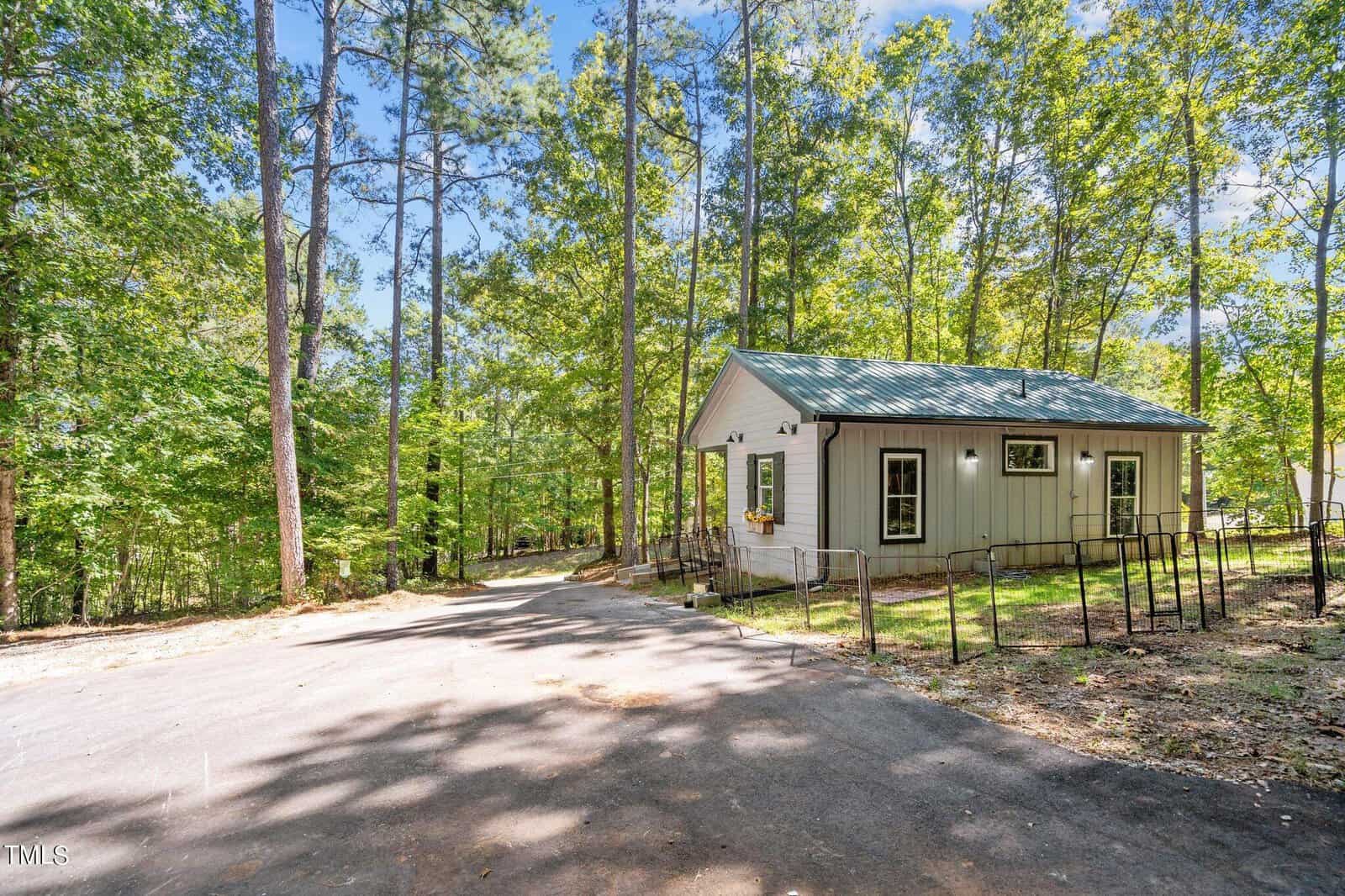 A small, light gray house with a green metal roof sits among tall trees. A paved driveway curves in front, and sunlight filters through the leafy forest creating a peaceful, wooded setting.