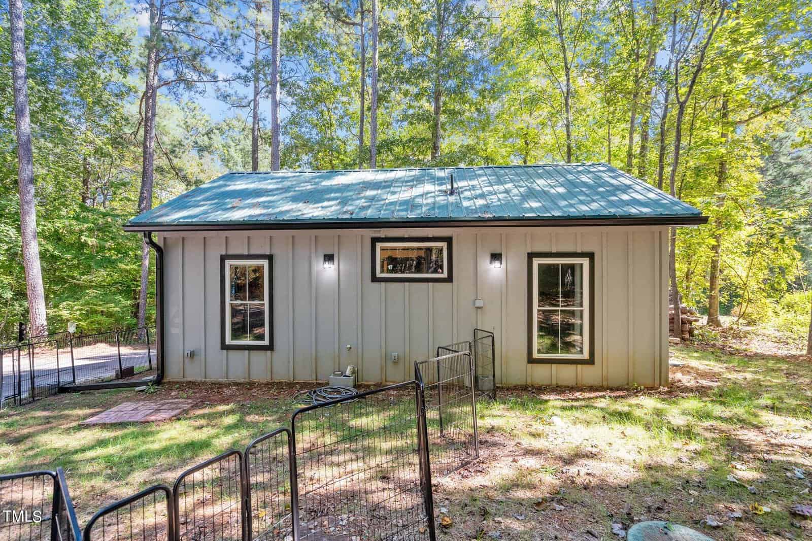 A small, single-story house with a metal roof and light-colored siding stands in a wooded area. Two windows and outdoor lights are visible, with a metal fence and trees surrounding the yard.